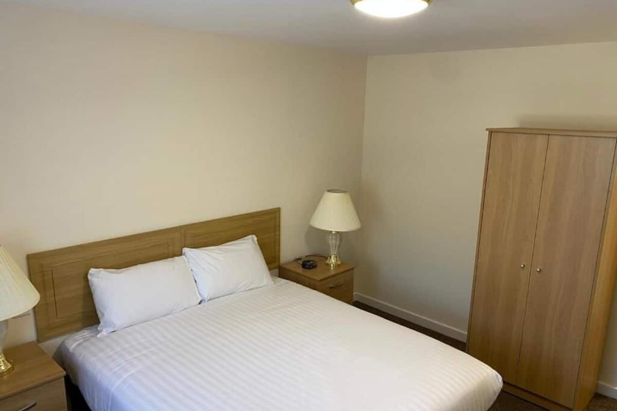 Uma cama bem arrumada com lençóis brancos, cabeceira de madeira, luminária de cabeceira e guarda-roupa.