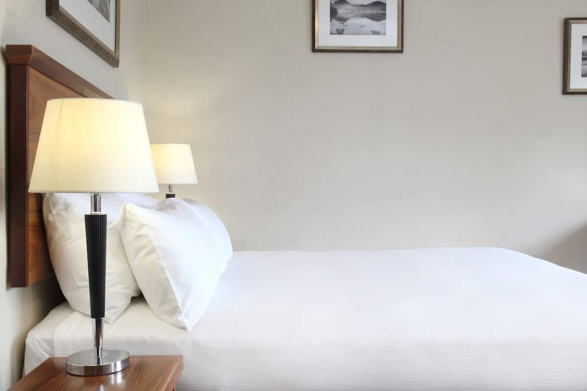 Uma cama bem arrumada com roupa de cama branca, duas luminárias de cabeceira e uma mesinha de cabeceira de madeira.