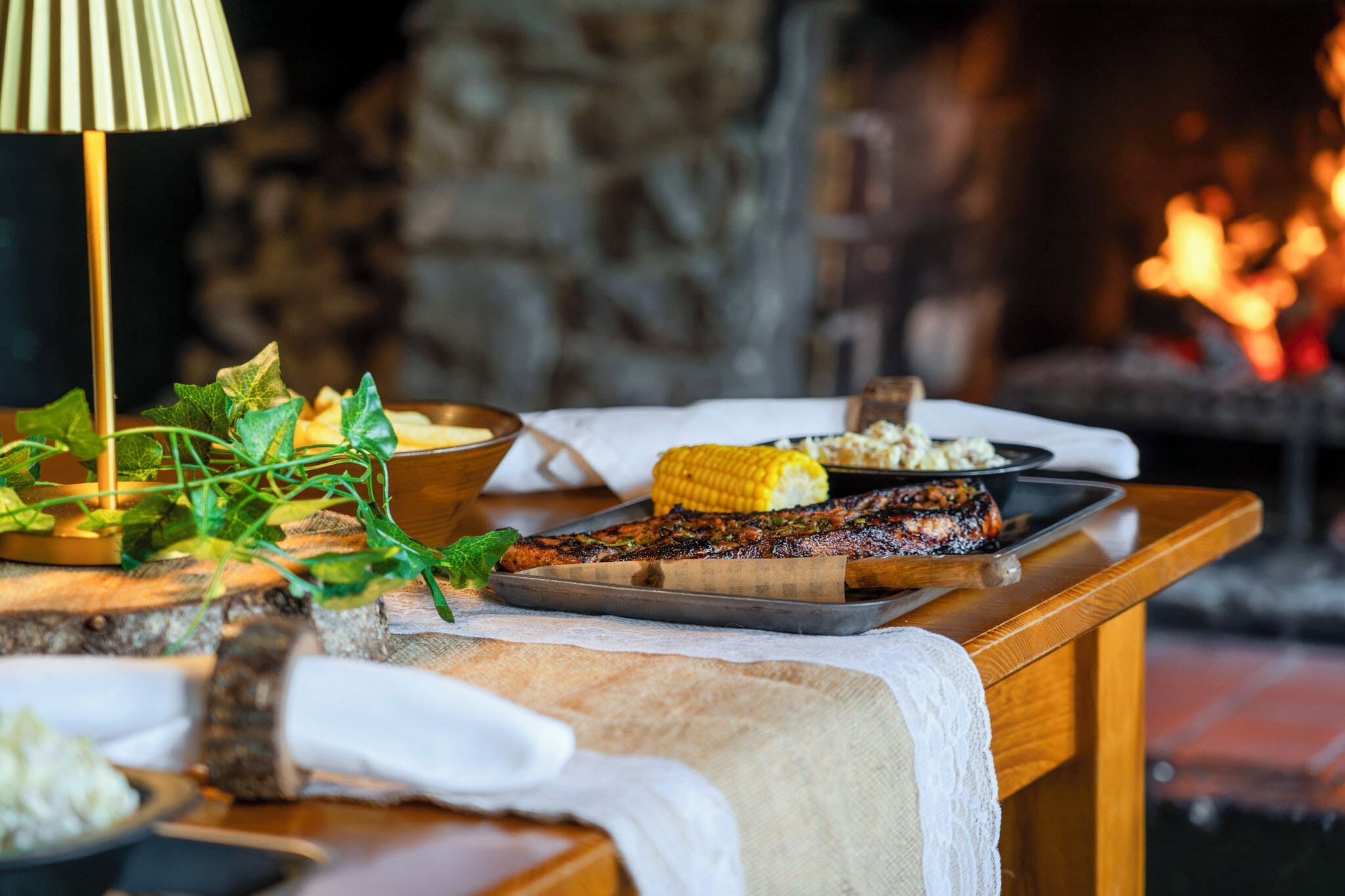 A wooden table with a tray of grilled meat, a bowl of corn, and a candle holder.
