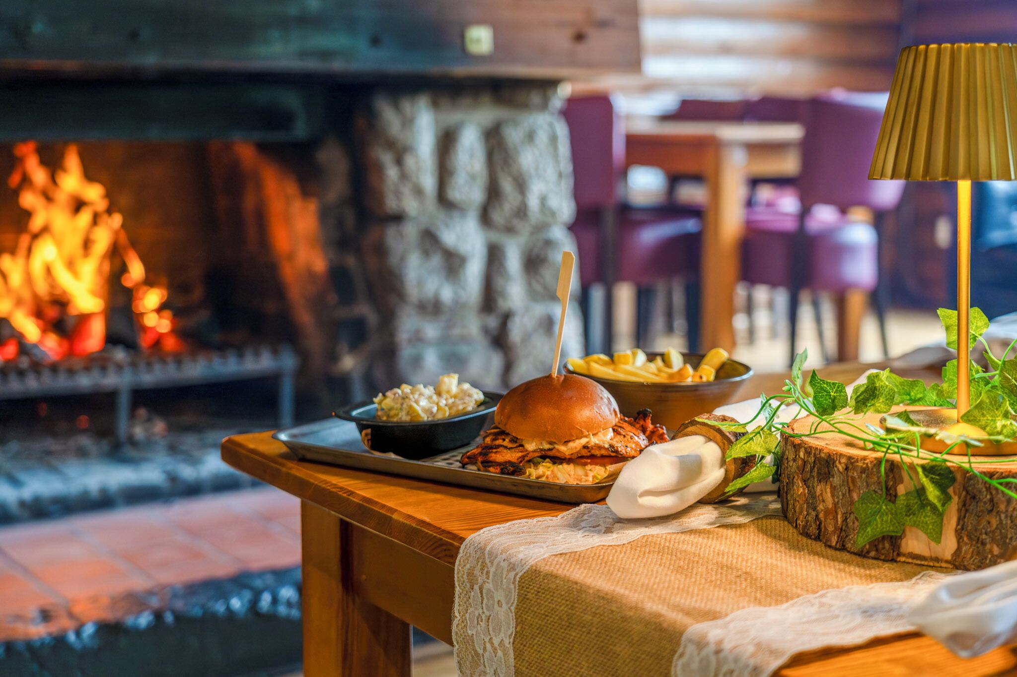 Una mesa de comedor con una hamburguesa, papas fritas y una ensalada, frente a una pared de piedra con chimenea.