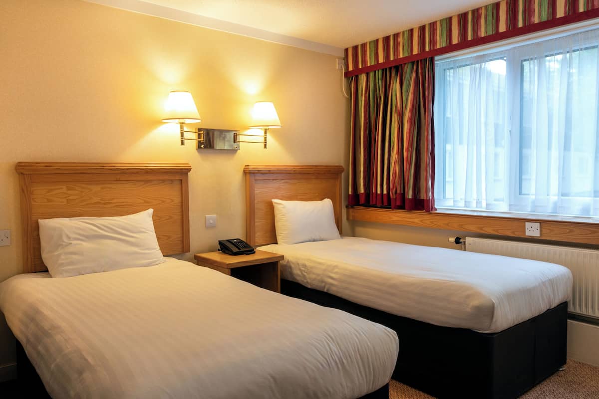 Quarto de hotel com duas camas, uma janela com cortinas e duas luminárias fixadas na parede.