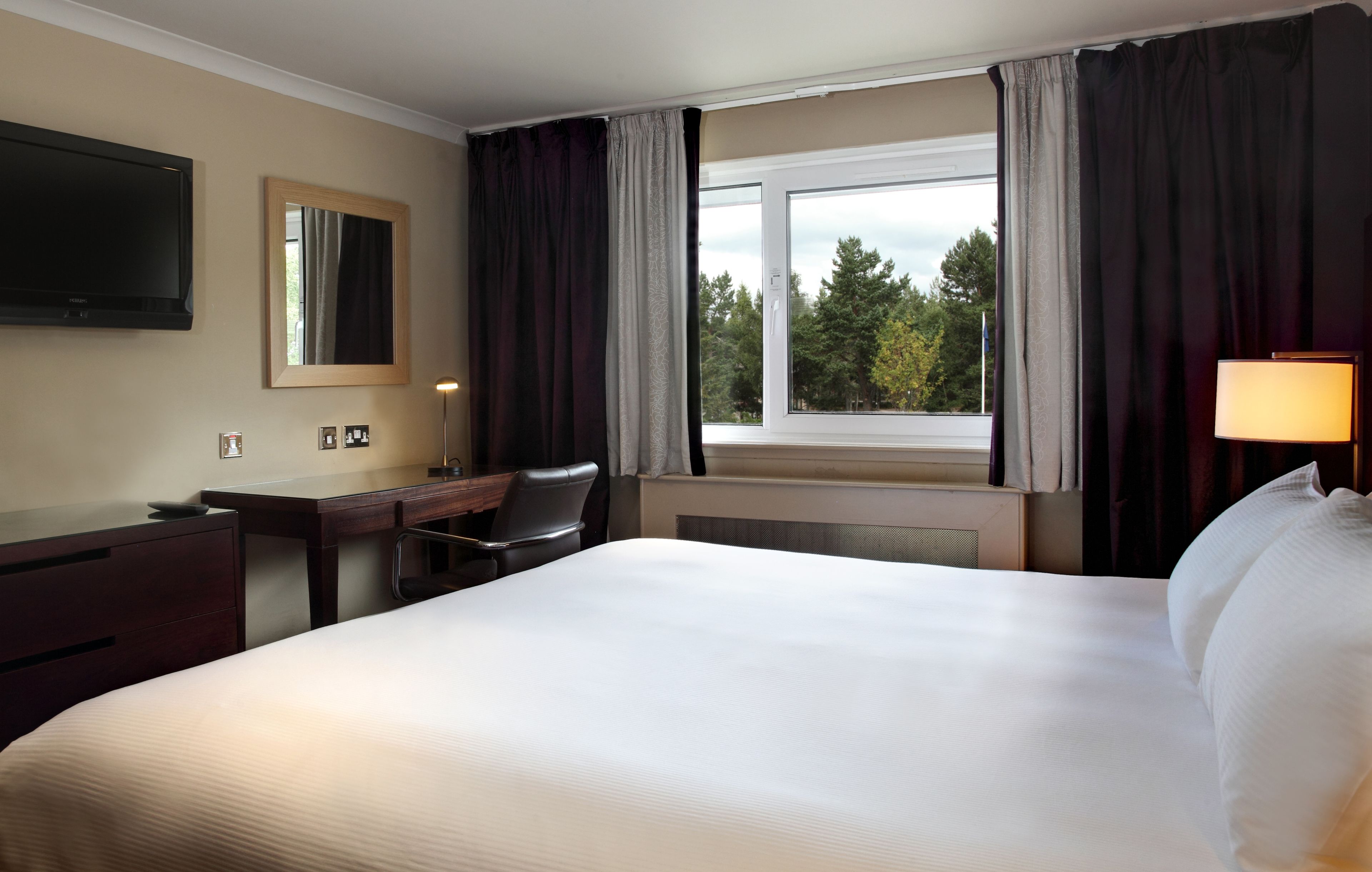 A hotel room with a large bed, a desk, a chair, a TV, and a window with curtains.