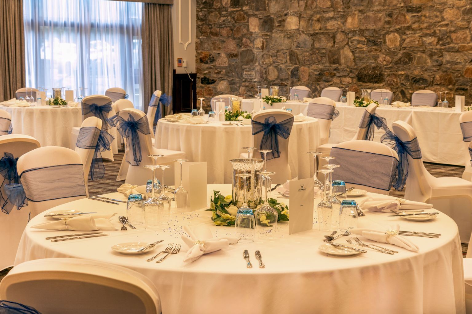A banquet hall with round tables set for a formal event, featuring white tablecloths, blue chair sashes, and floral centerpieces.