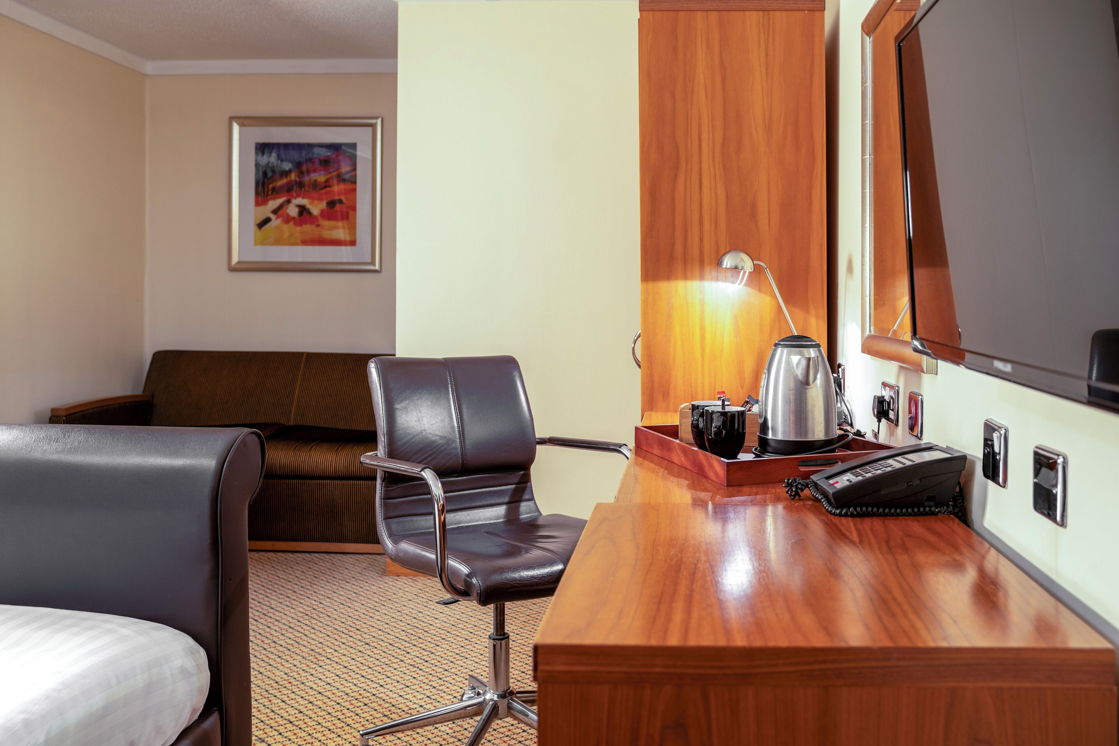 A hotel room with a wooden desk, a black leather chair, a bed, a sofa, a painting on the wall, a lamp, and a telephone.
