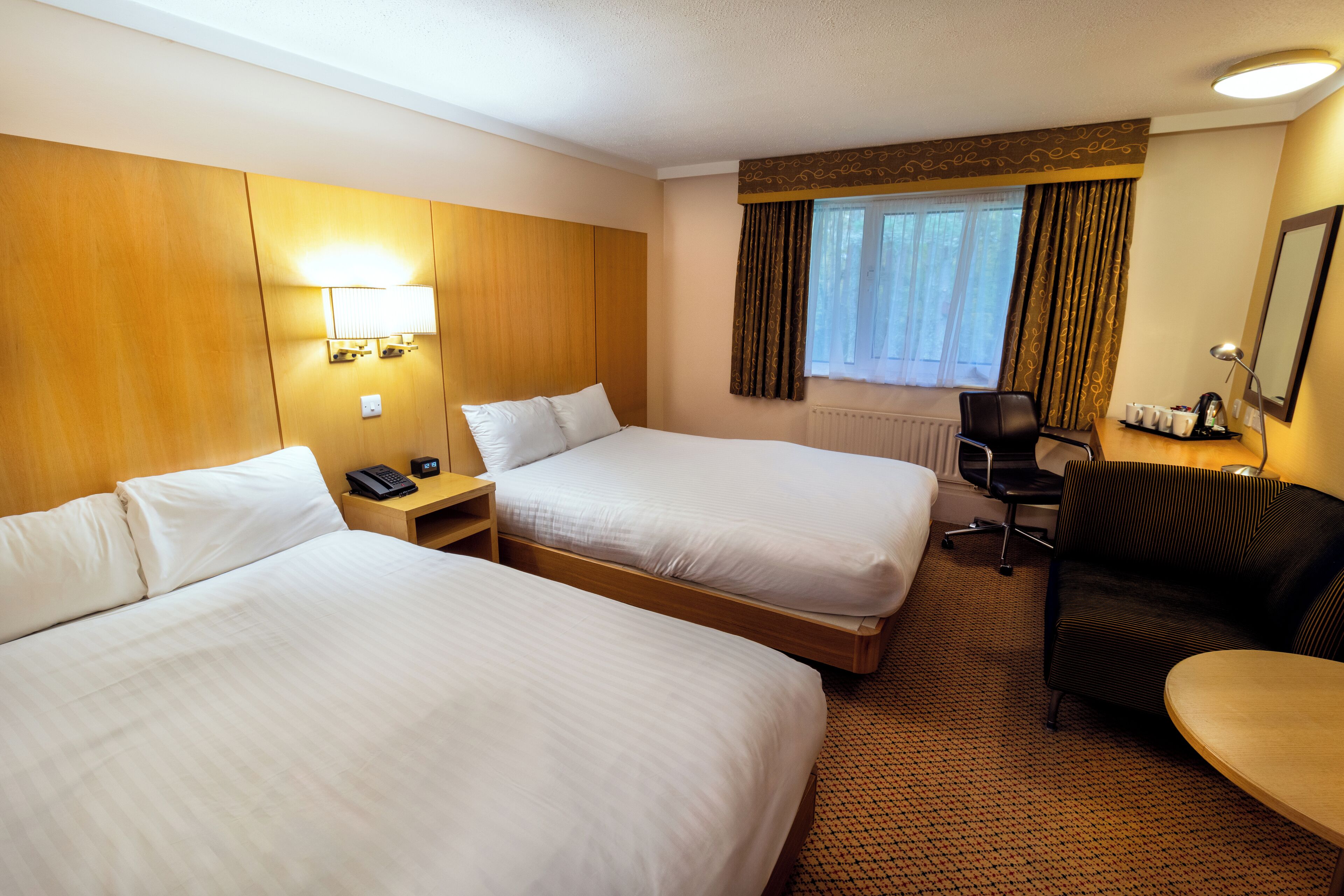 A hotel room with two beds, a desk, a chair, a lamp, and a window with curtains.