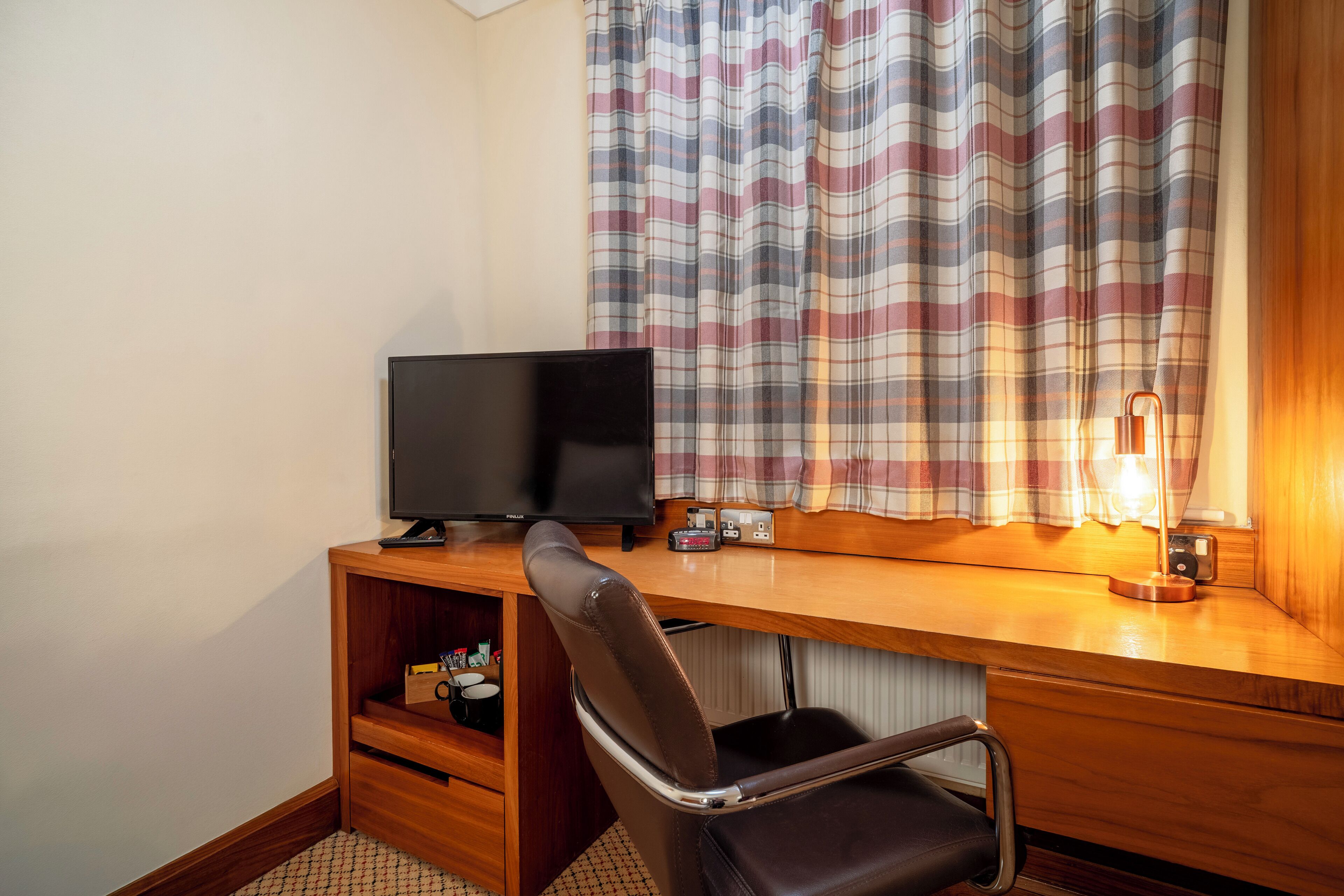 Un escritorio de madera con una televisión, una silla, una lámpara y una ventana con cortinas a cuadros.