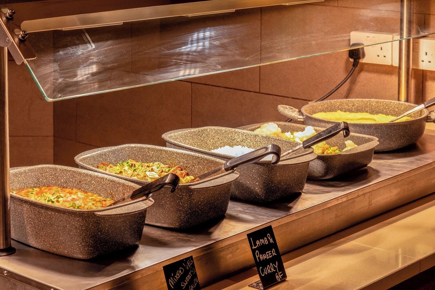 Un mostrador de buffet con diversas opciones de comida, incluyendo verduras mixtas, curry de paneer y otros platos.