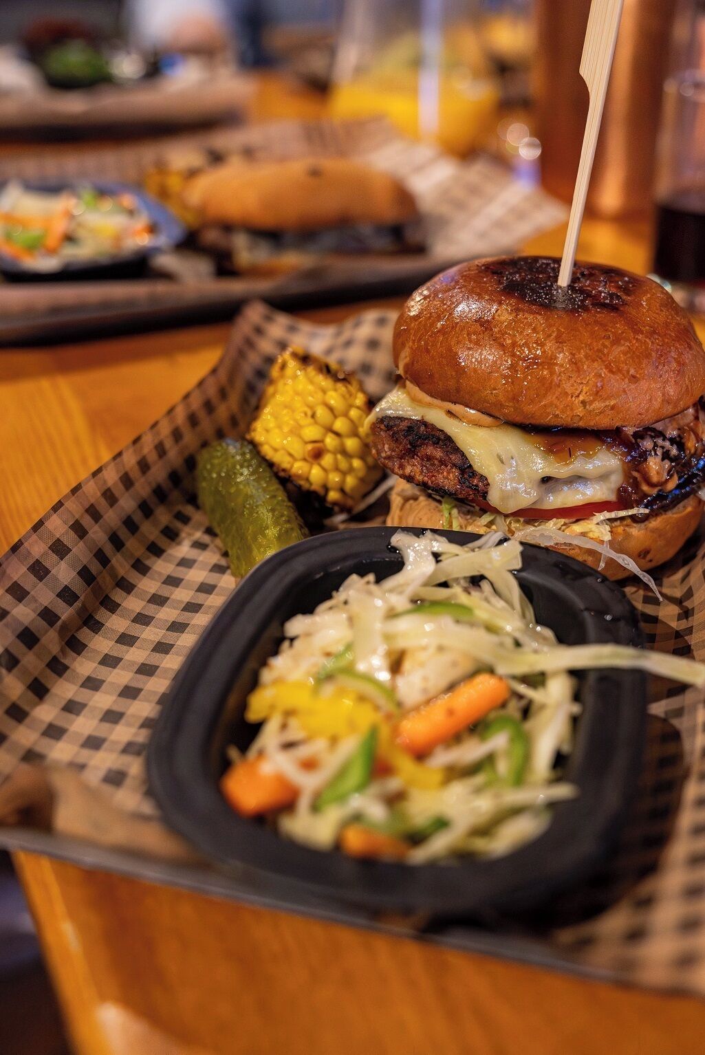 Een hamburger met een satéprikker, een portie koolsalade en een augurkje op een geruit kartonbakje.