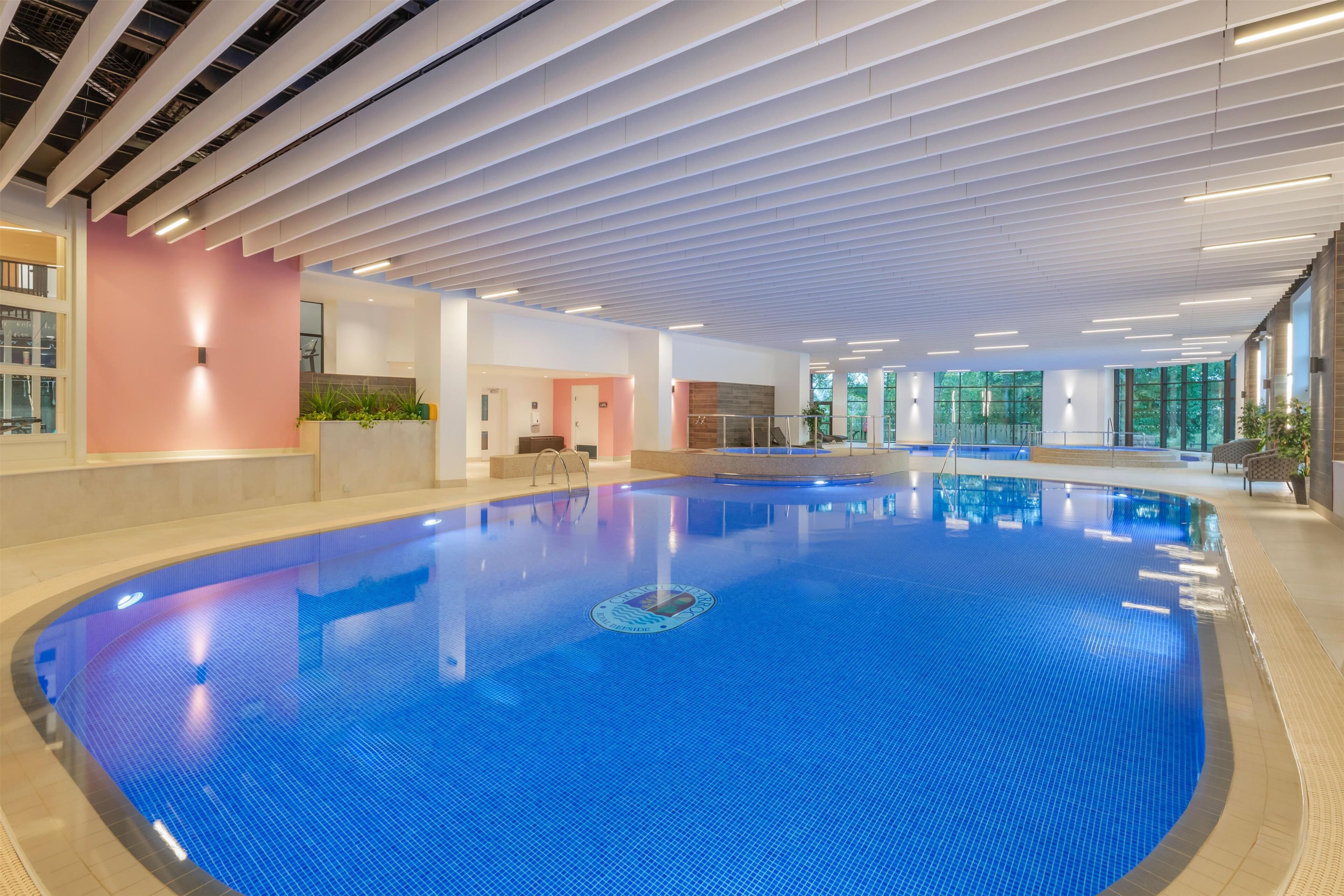 An indoor swimming pool with a blue tiled floor, surrounded by walls with a pink accent wall and large windows allowing natural light.
