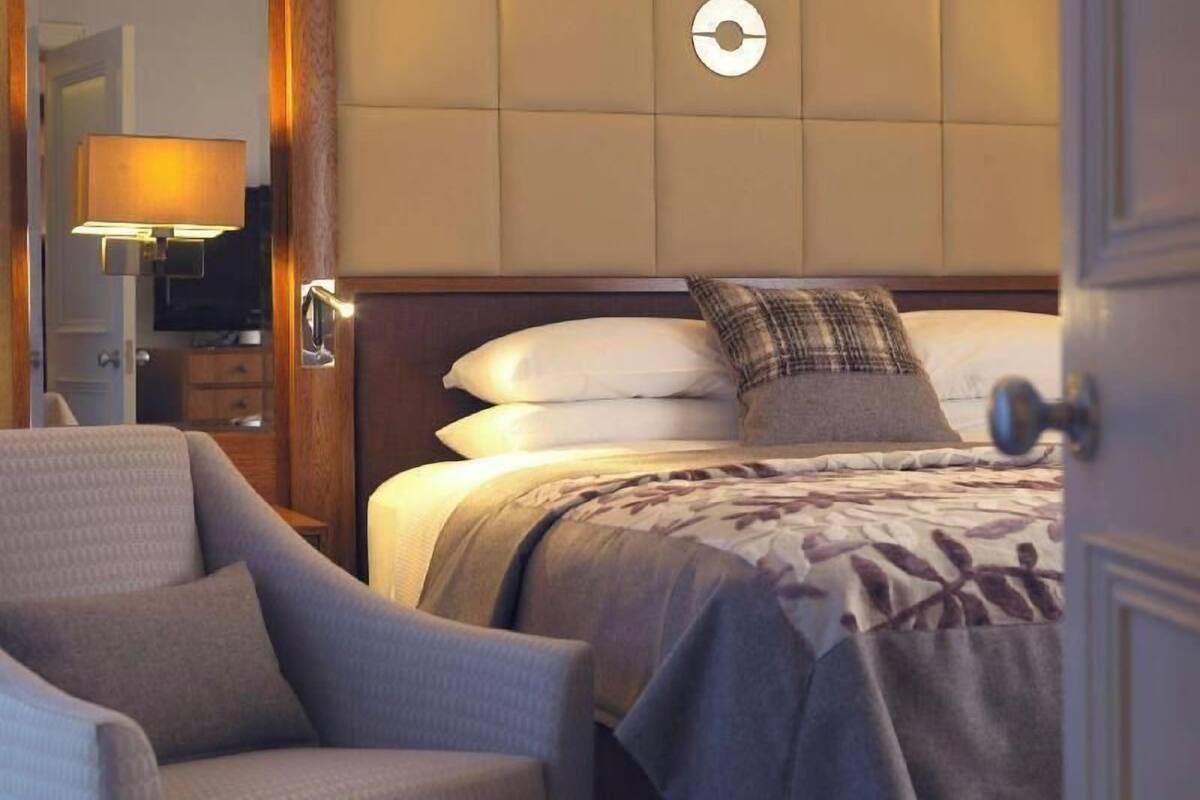 A hotel room with a bed, a chair, a bedside table, a lamp, and a wall clock.