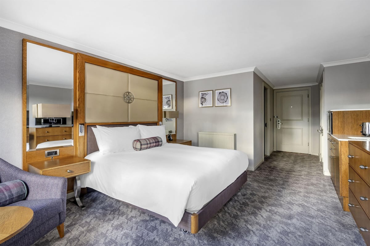 A hotel room with a large bed, a wooden nightstand, a chair, a dresser, and a large mirror.