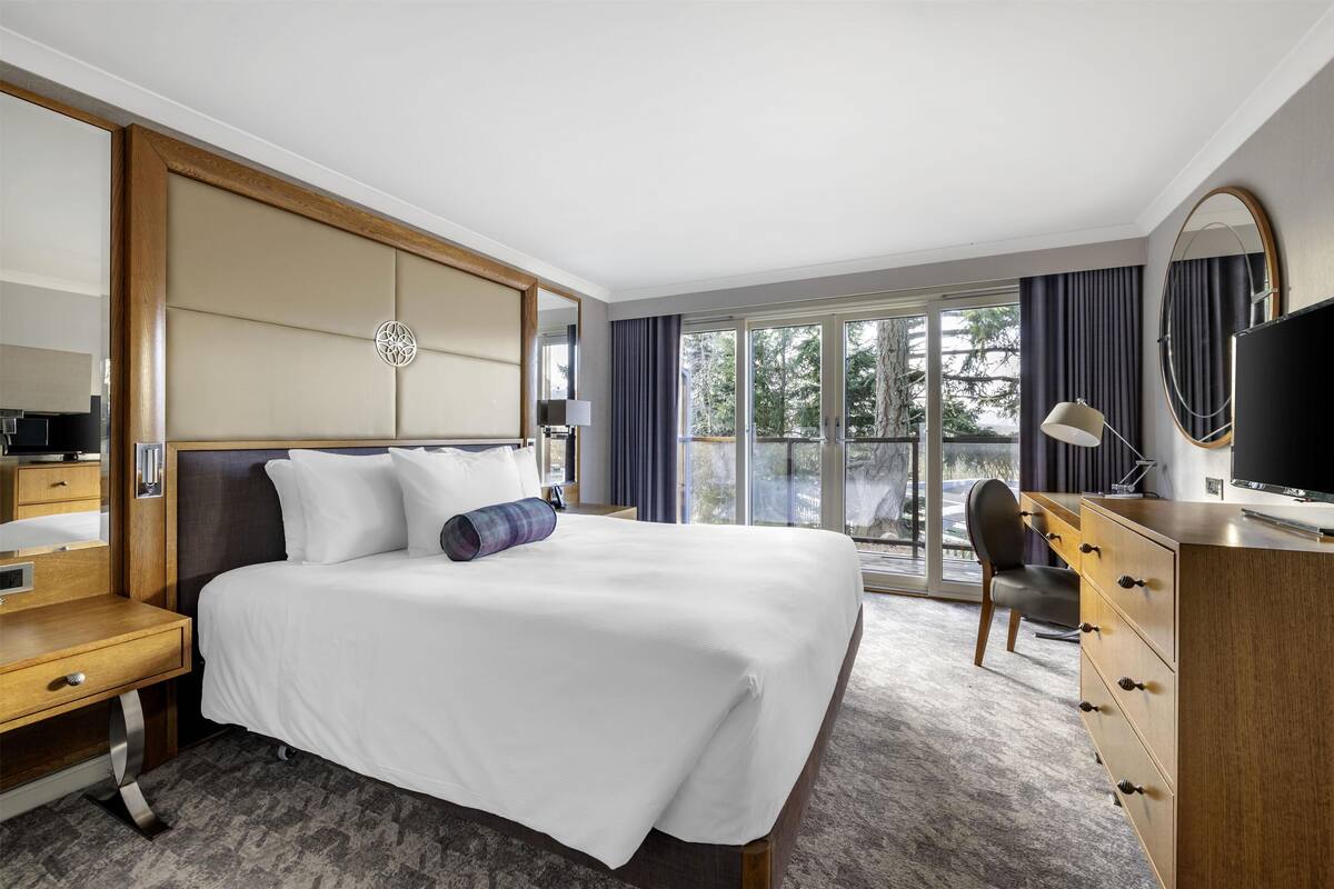 A modern hotel room with a large bed, a wooden desk, a dresser, a TV, and a balcony with a view of trees.