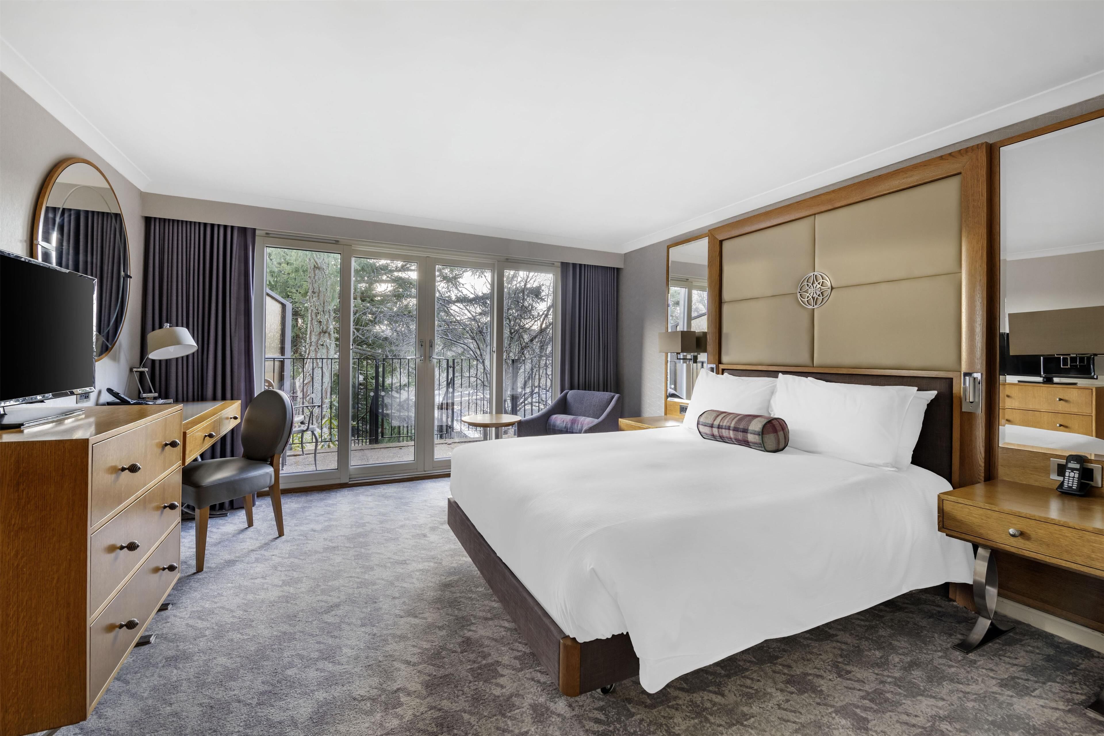 A hotel room with a large bed, a desk with a computer, a chair, a dresser, and a large window with a view of trees.