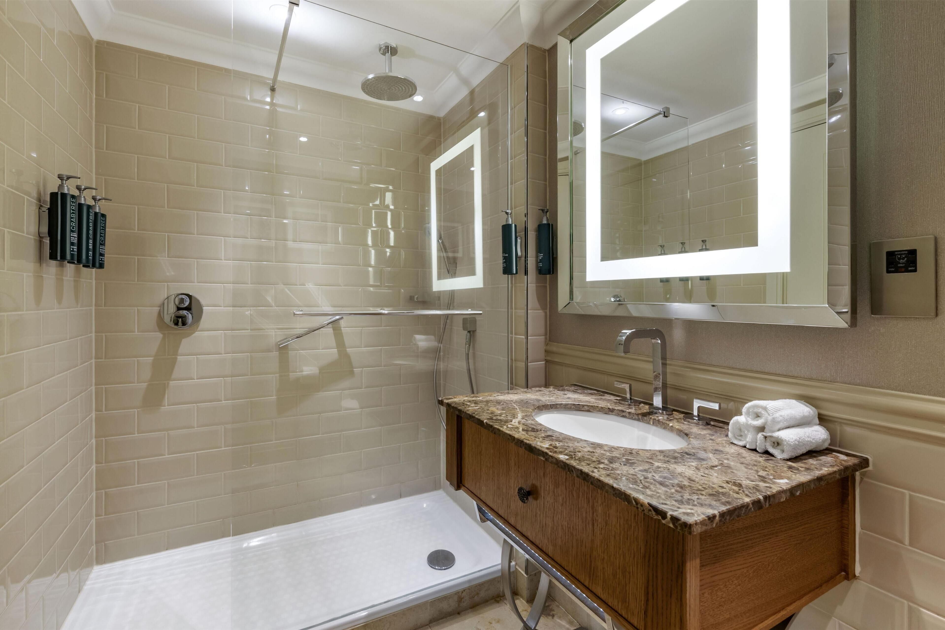 A modern bathroom with beige tiles, a glass shower enclosure, a wooden vanity with a sink, and a large mirror.