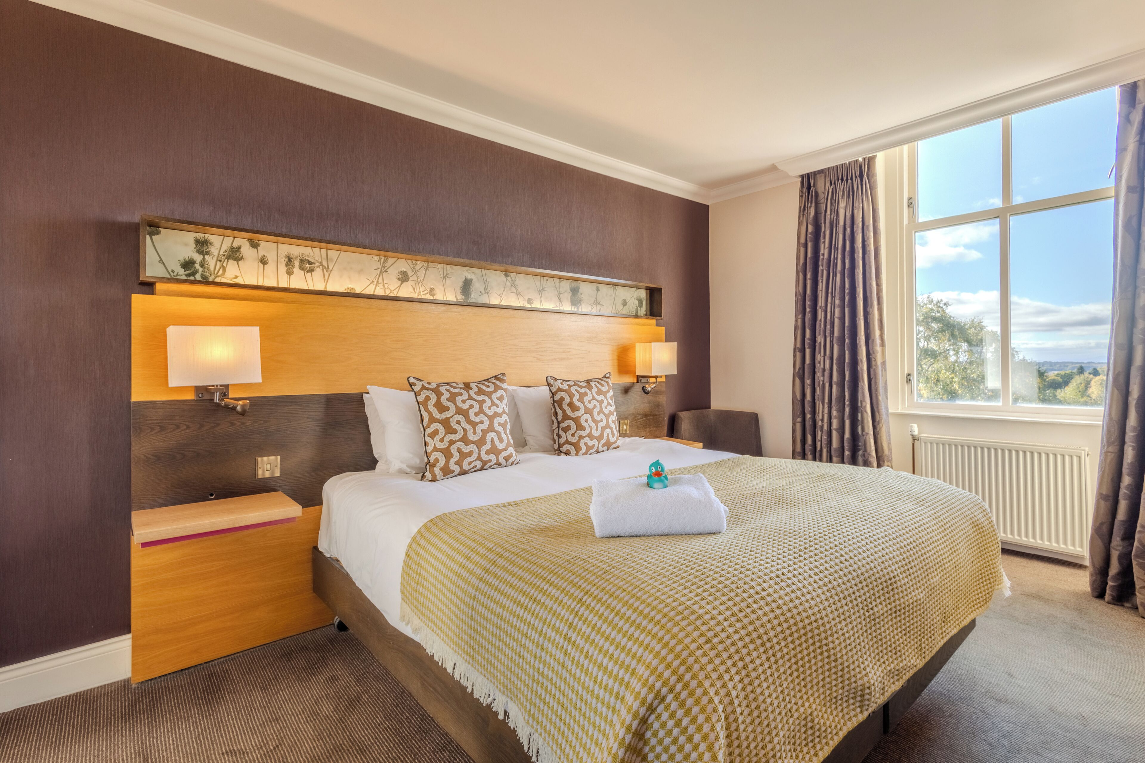 A hotel room with a large bed, two bedside lamps, a headboard with a floral design, a view of the outdoors through a window, and a neatly made bed with a towel.