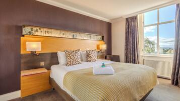 Habitación de hotel con una cama grande, dos lámparas de noche, un cabecero con diseño floral, vistas al exterior por una ventana, y una cama bien hecha con una toalla.