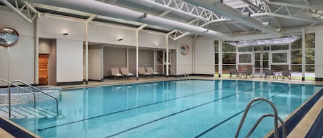 Una piscina interior con agua cristalina y azul, rodeada de sillones de descanso y un techo alto con tragaluces.