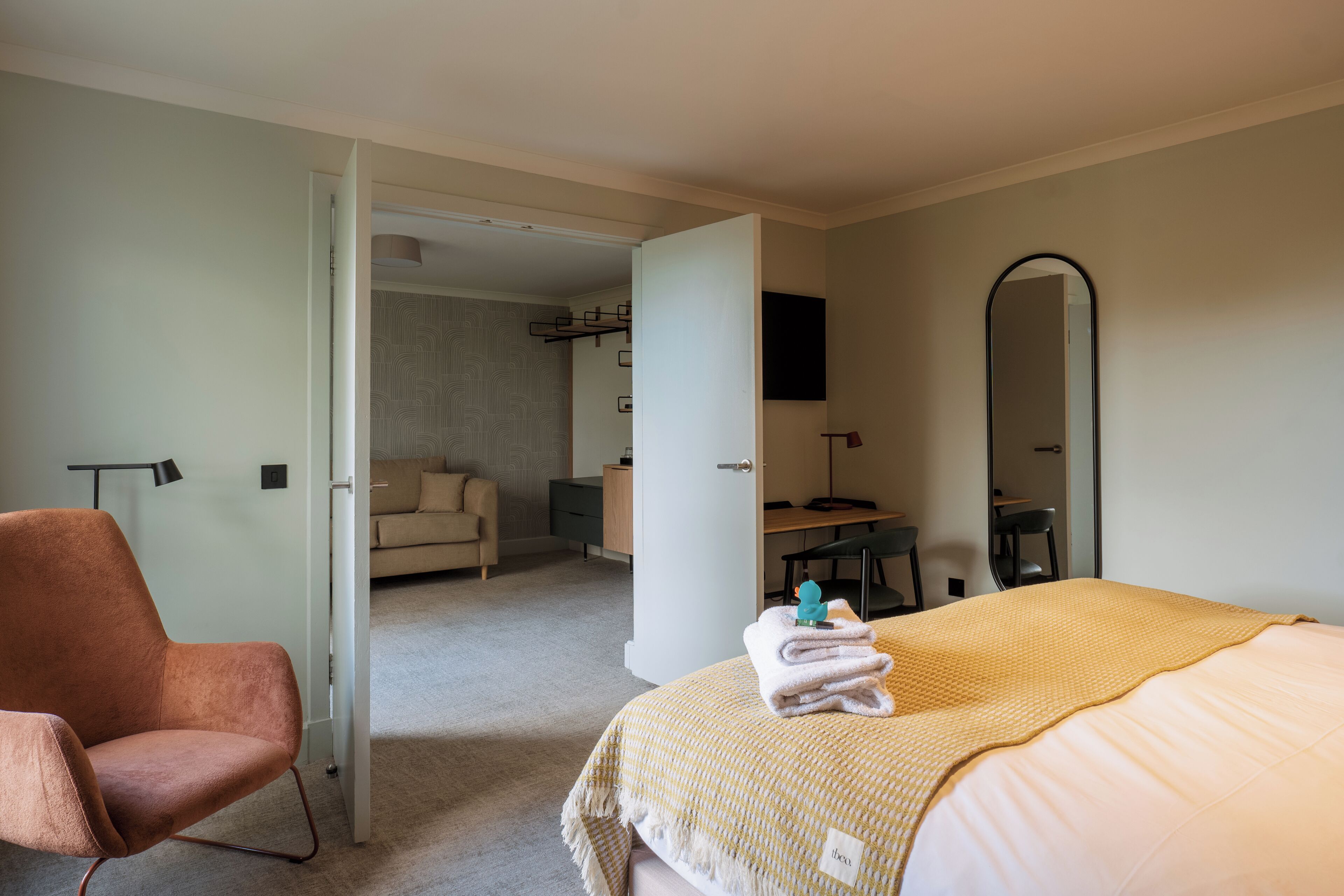 A hotel room with a bed, a chair, a desk, a mirror, and a door leading to another room.