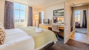 A hotel room with a bed, desk, and computer. There is a window with curtains, a lamp, and a painting on the wall.