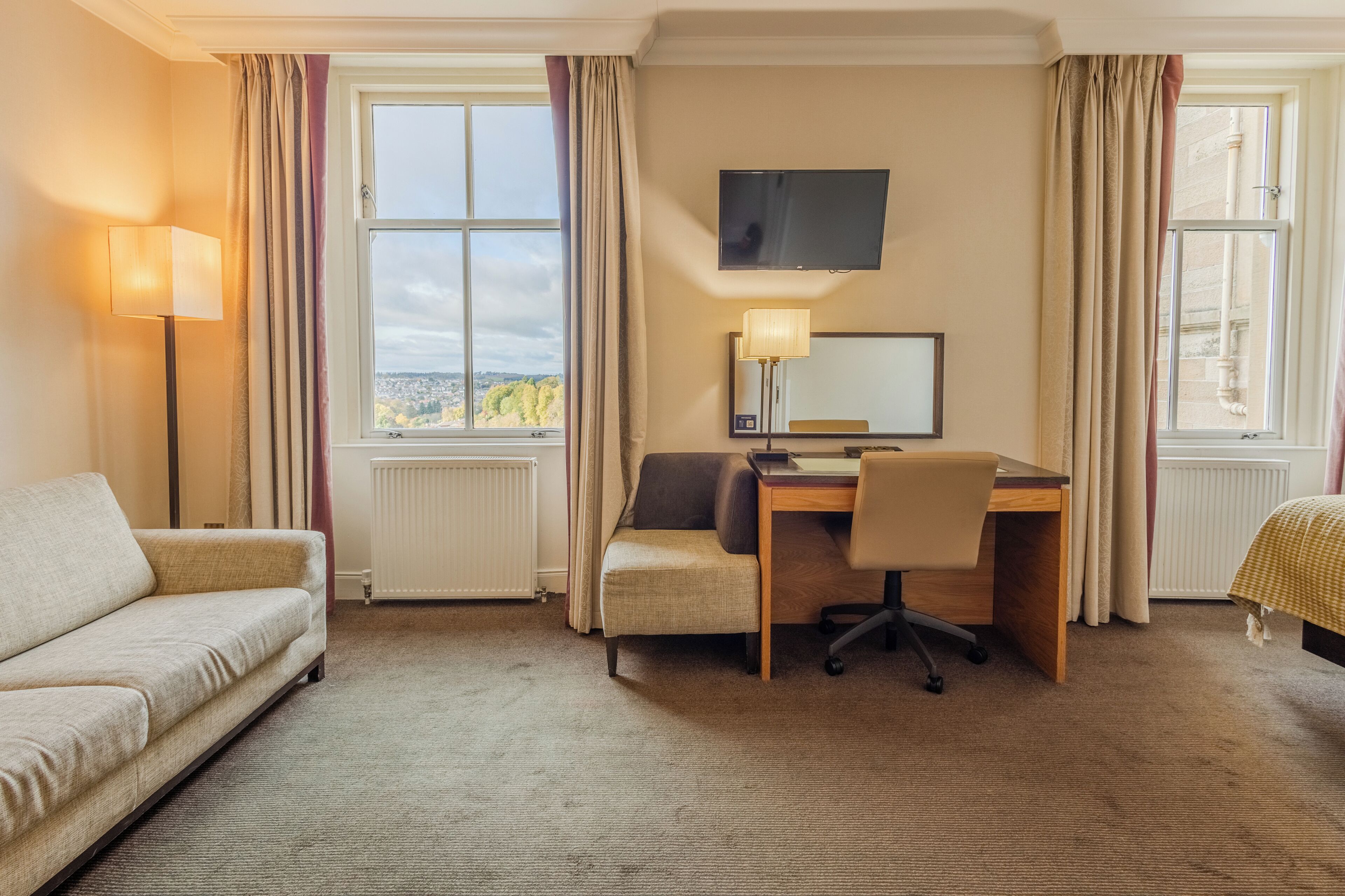 A hotel room with a sofa, a desk with a chair, a television, and a window with curtains.