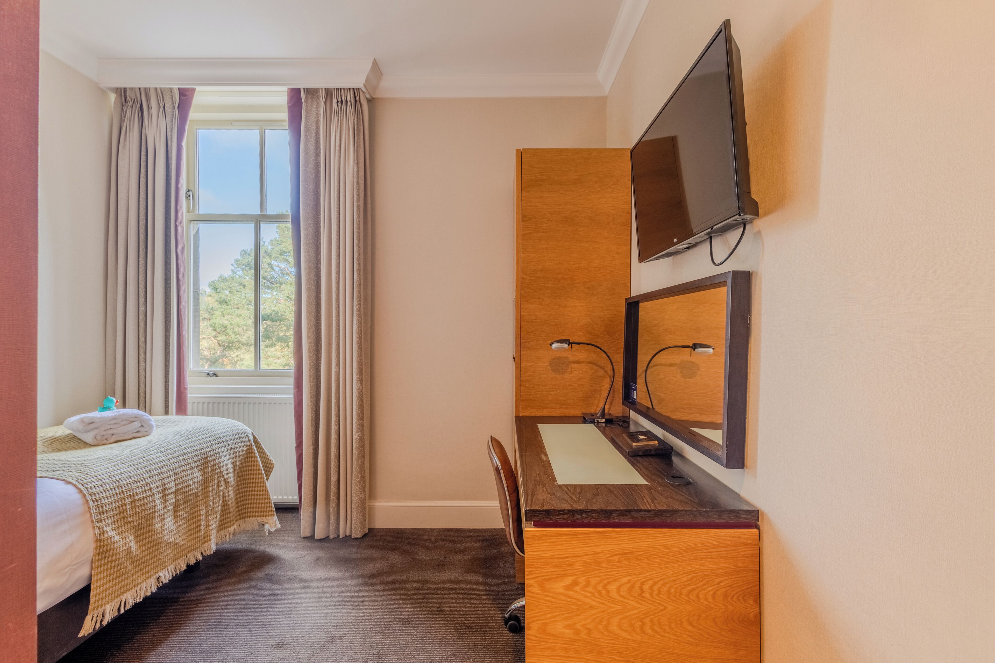 A hotel room with a bed, a desk with a computer, a TV, and a window with curtains.