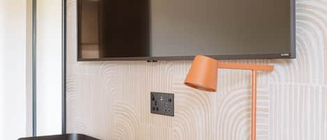 A modern room with a flat-screen TV mounted on the wall, a wooden desk with a remote control, and an orange floor lamp.