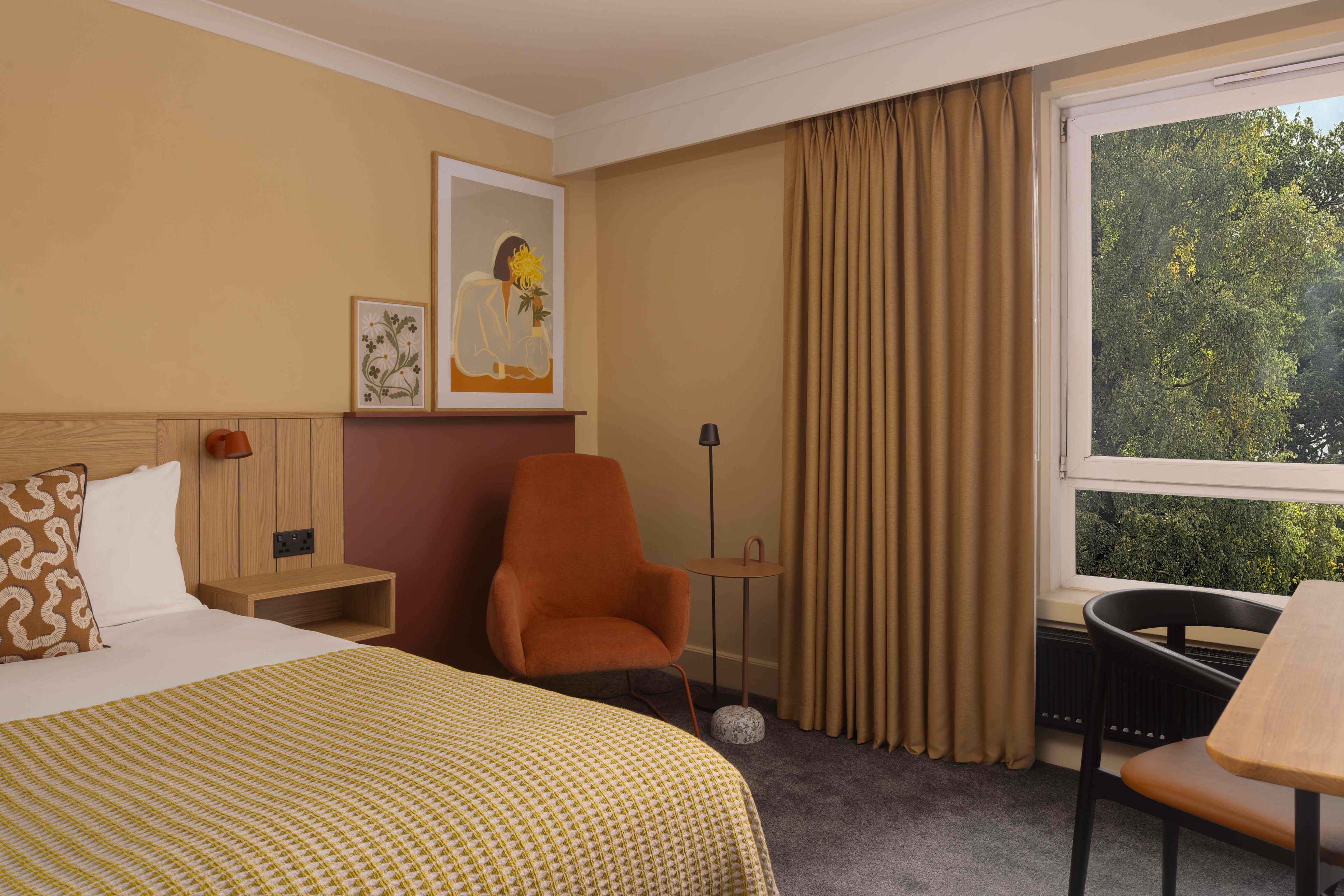 A hotel room with a bed, a chair, a desk, a window with curtains, and artwork on the wall.
