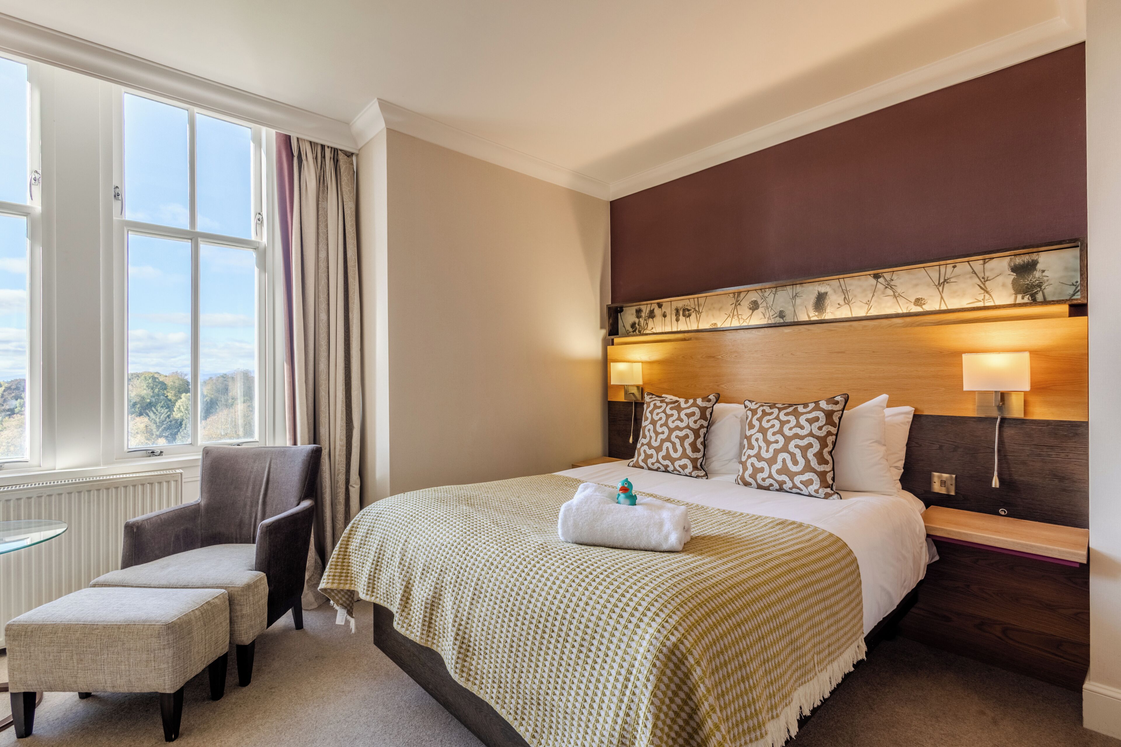 A hotel room with a large bed, a chair, a small table, and a view of the outdoors.