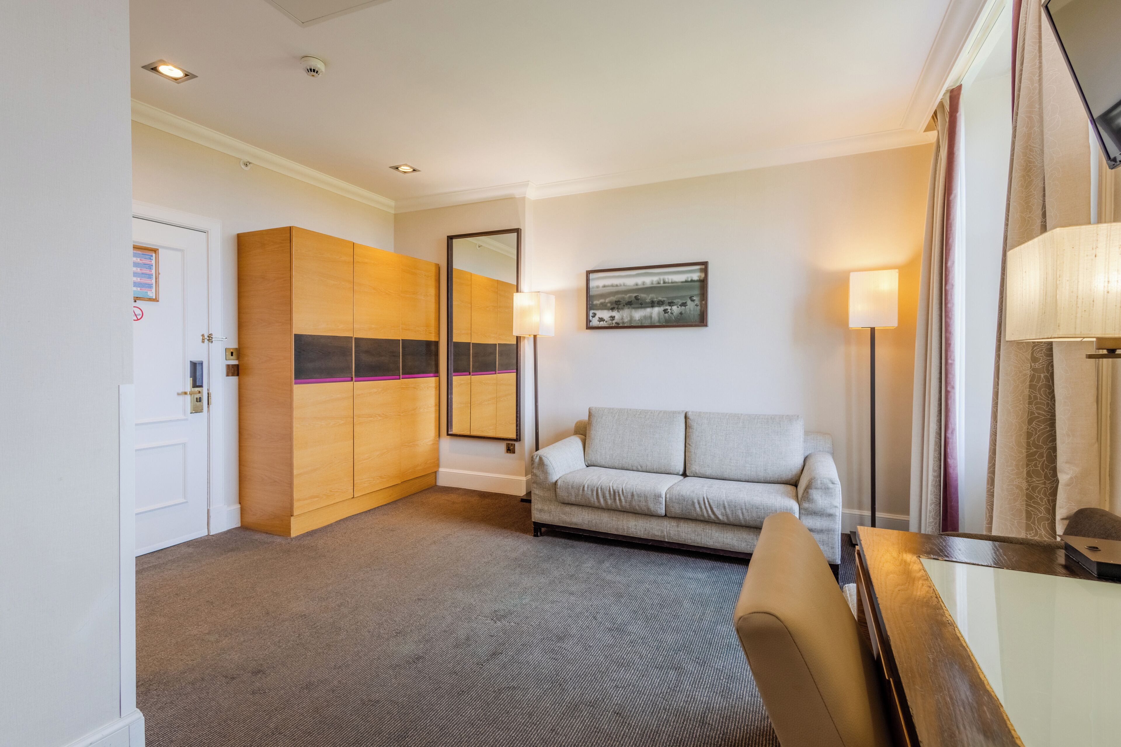 Une chambre d’hôtel avec un canapé gris, une armoire en bois, un miroir, un tableau encadré au mur et une lampe.