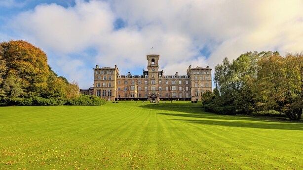 A large, historic building with a central tower and multiple wings, surrounded by a well-maintained lawn and trees.