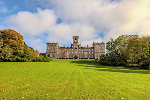 A large, historic building with a central tower and multiple wings, surrounded by a well-maintained lawn and trees.