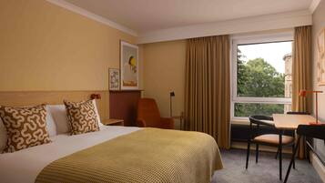 Habitación de hotel con cama, escritorio, silla, ventana con cortinas y un cuadro en la pared.