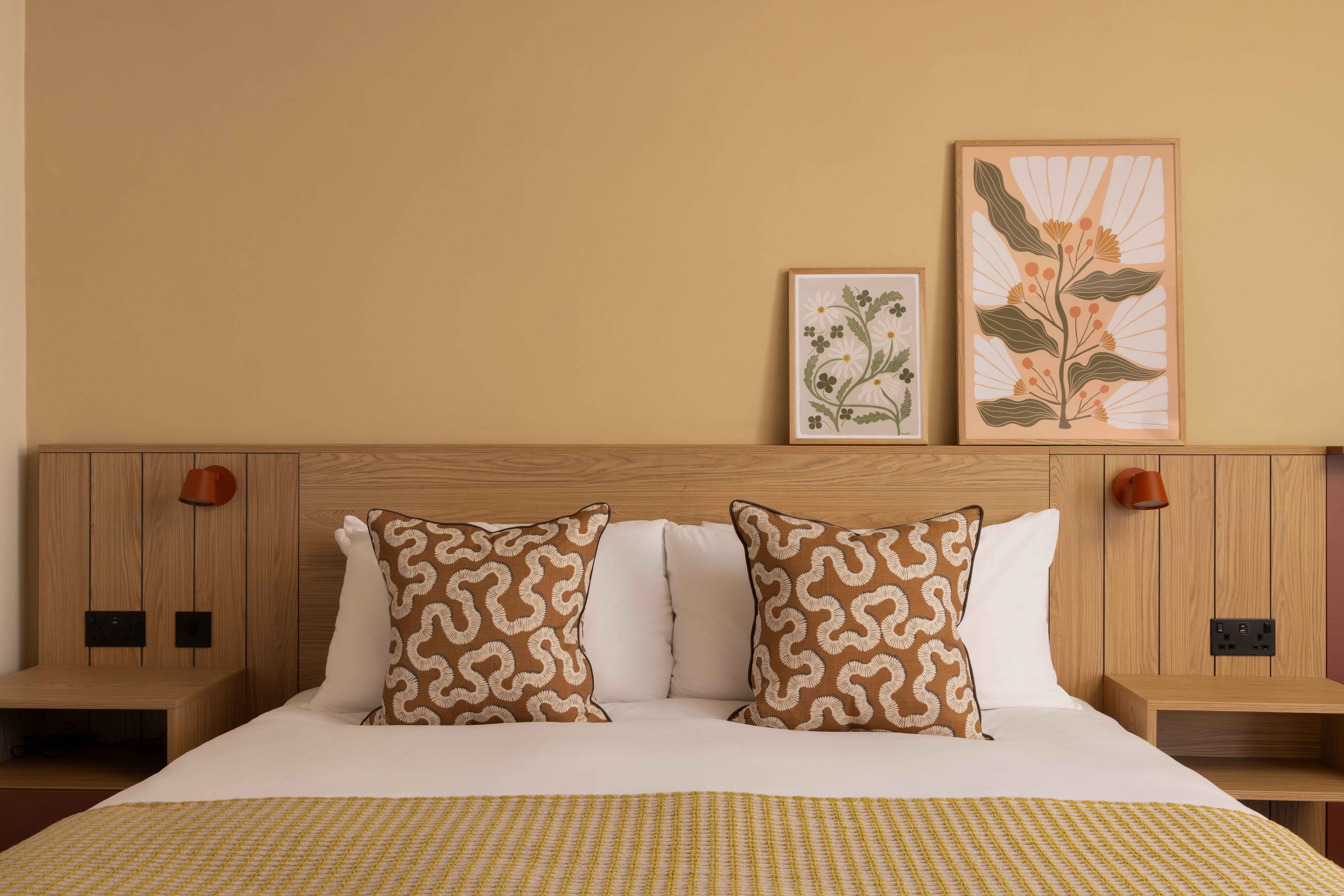 A hotel room with a wooden headboard, two bedside tables, a bed with white bedding, patterned pillows, and two framed floral artworks on the wall.