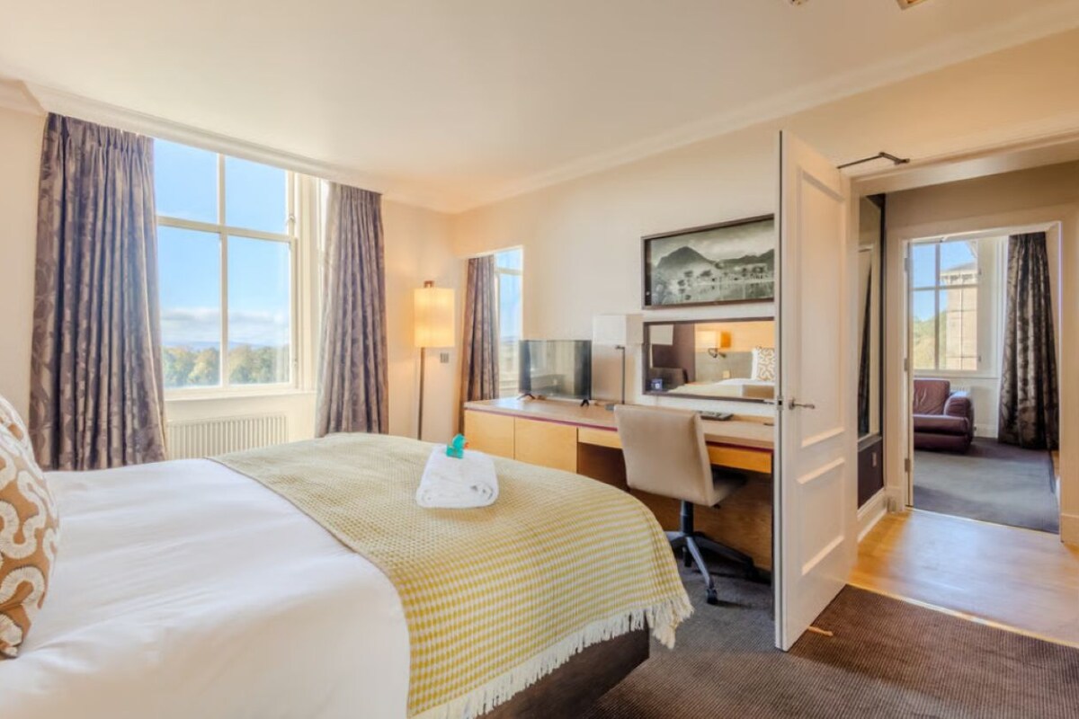 A hotel room with a bed, desk, and computer. There is a window with curtains, a painting on the wall, and a view of the outside.