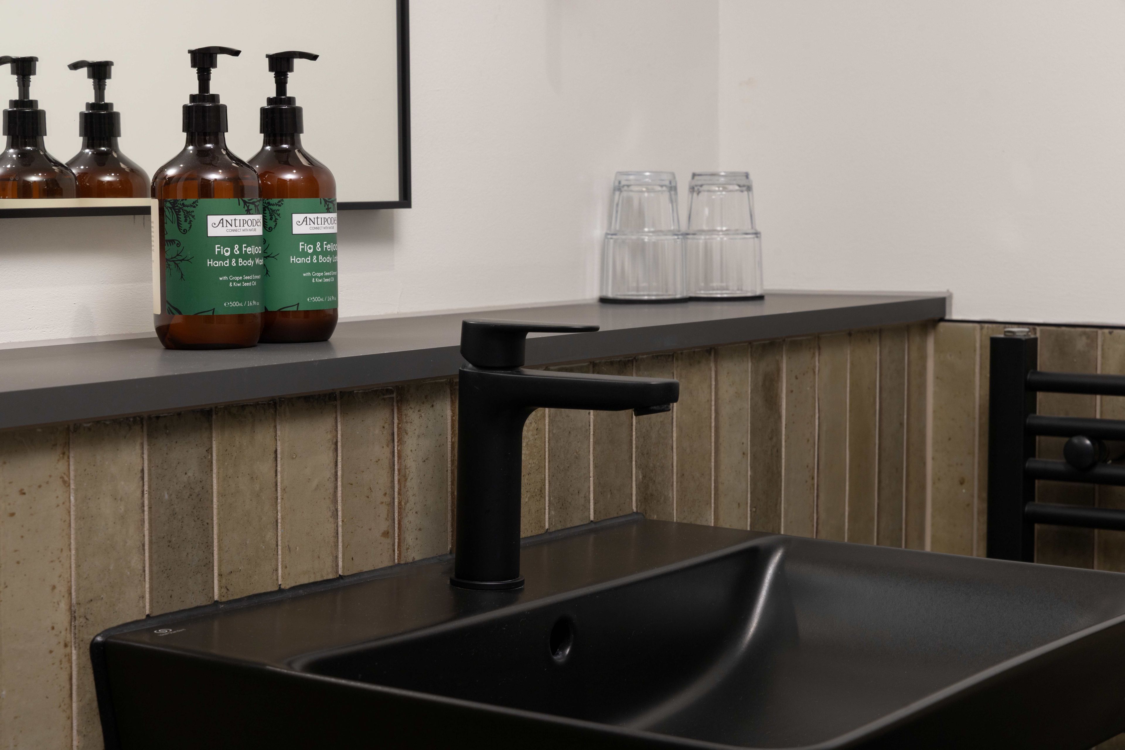 Un baño con un lavamanos negro, un espejo y una repisa con botellas y vasos.