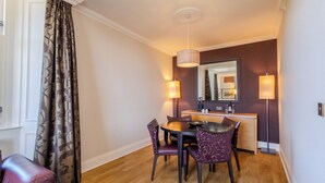 A hotel room with a dining area featuring a round table, purple upholstered chairs, and a wooden floor. There is a large mirror above the sink, and two wall-mounted lamps provide lighting.