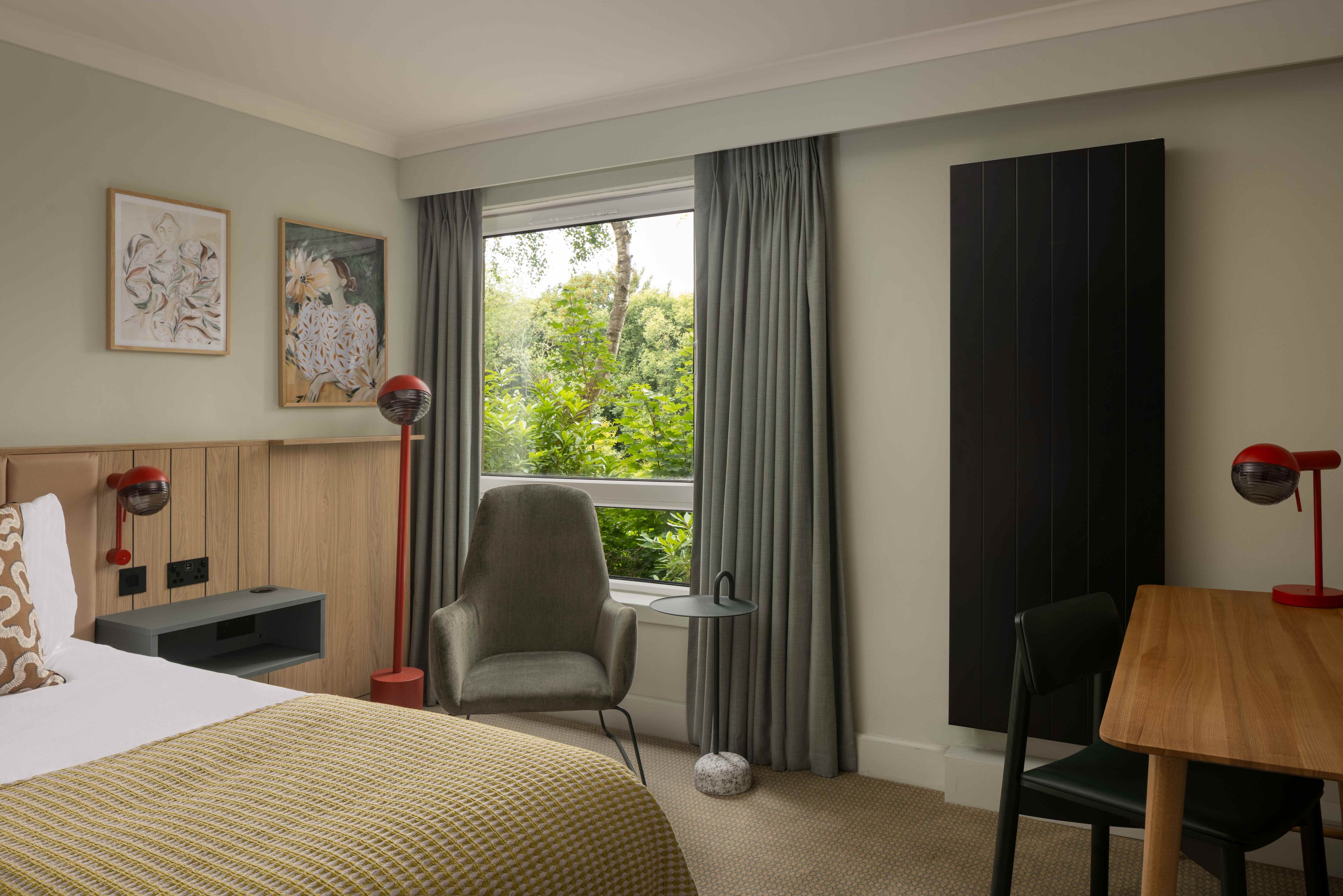 A hotel room with a bed, a desk, a chair, a window with curtains, and artwork on the wall.