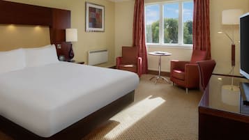 A hotel room with a large bed, two armchairs, a small table, a TV, and a window with curtains.