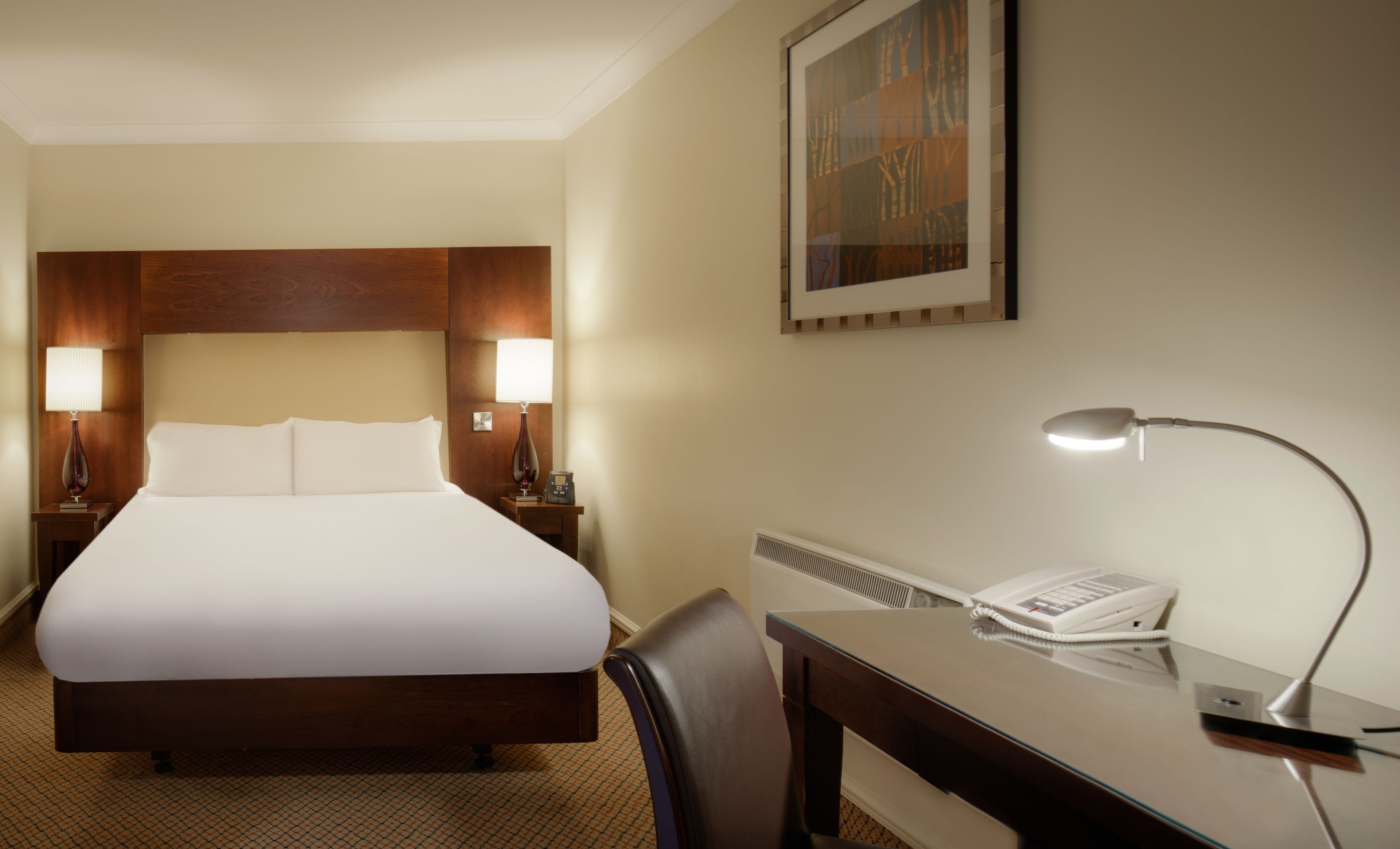 A hotel room with a large bed, two bedside lamps, a desk with a telephone, and a painting on the wall.