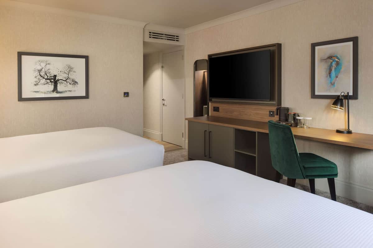 A hotel room with two beds, a desk, a chair, a TV, and artwork on the walls.