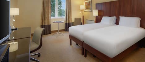 A hotel room with a large bed, a desk with a lamp, a chair, and a window with curtains.