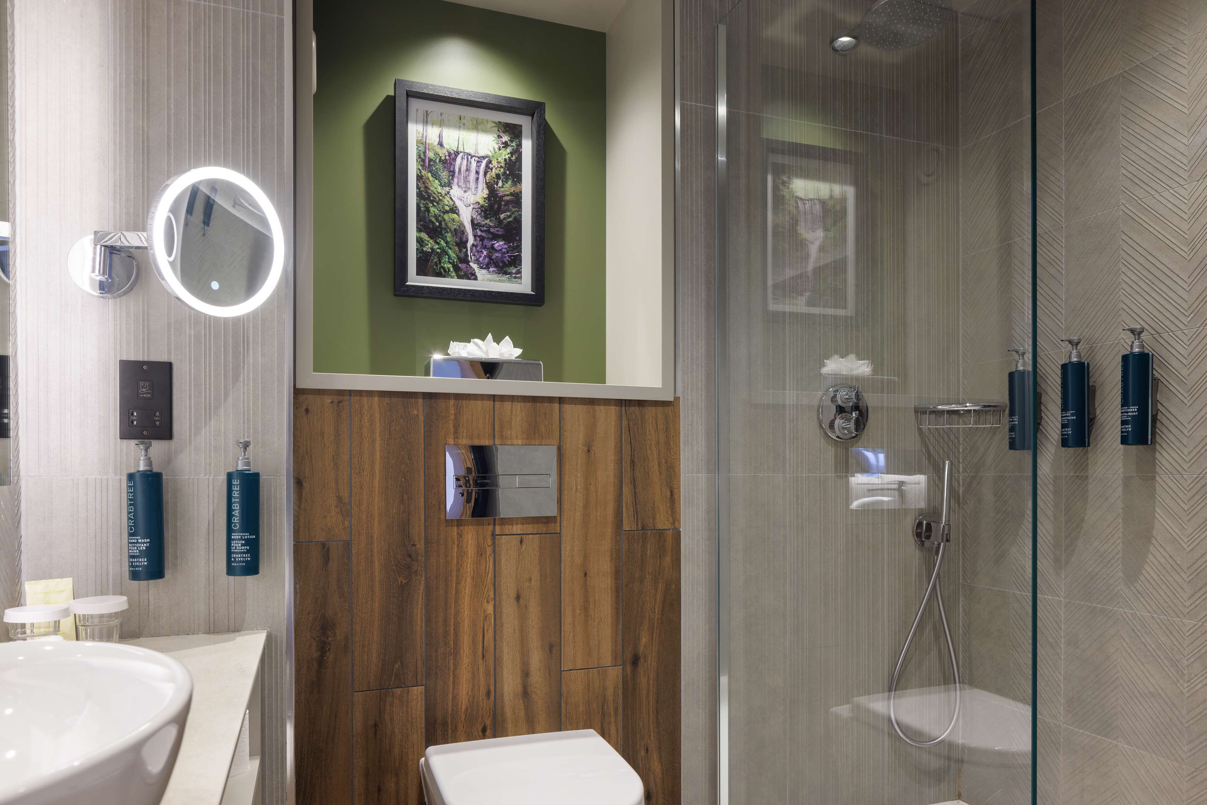 A modern bathroom with a wooden vanity, a round mirror, a framed picture, and a glass-enclosed shower area.
