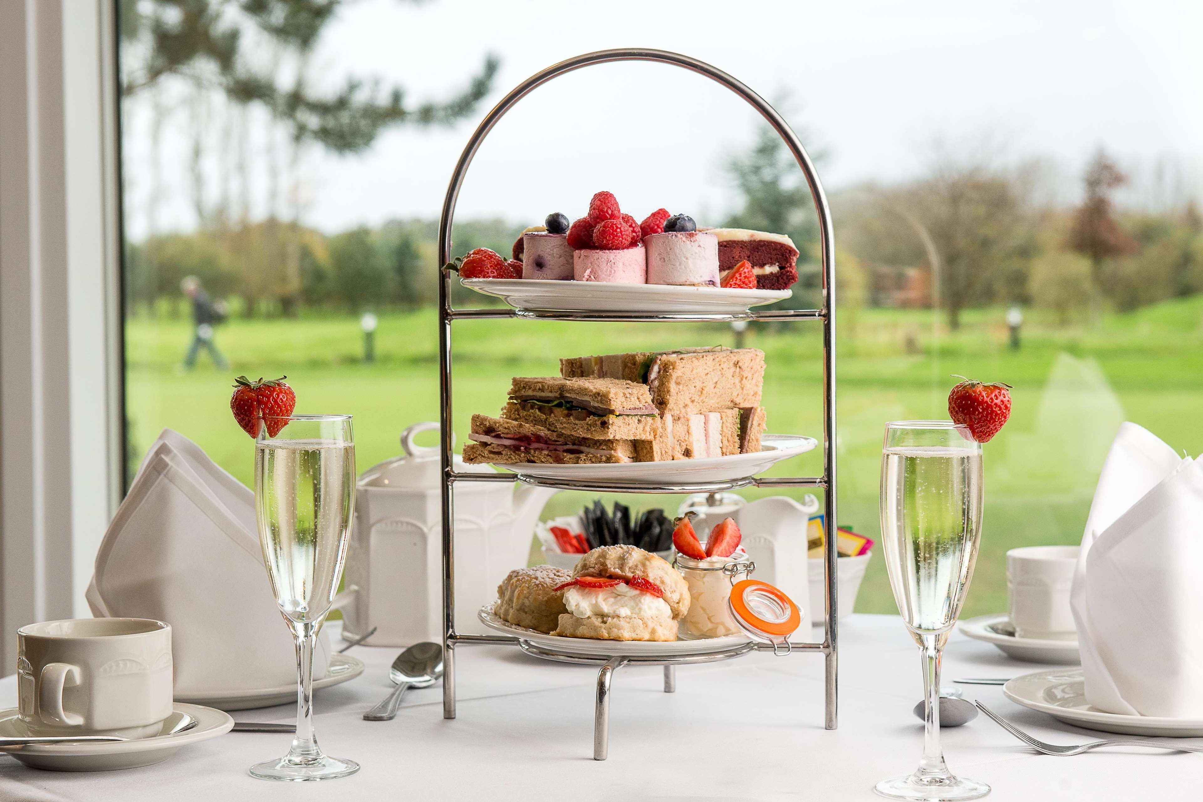 A three-tiered dessert stand with various pastries and a glass of champagne, set on a table with a view of a green landscape.
