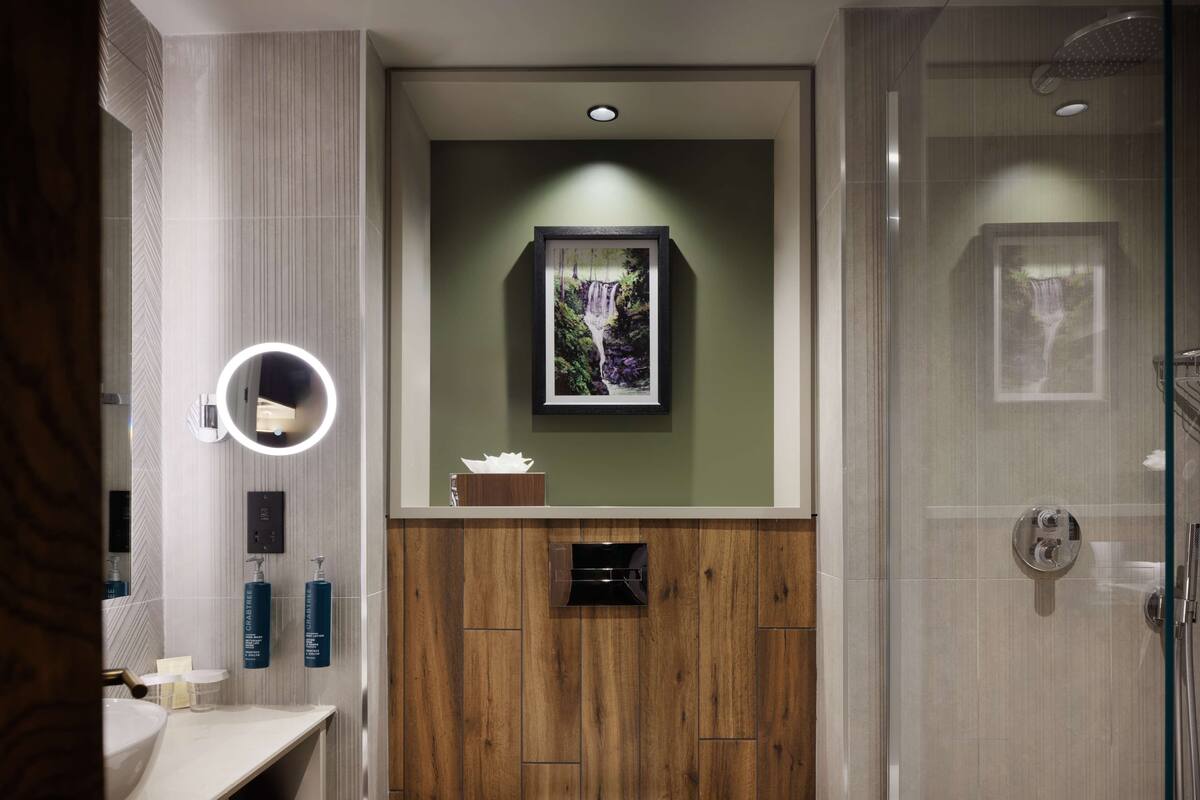 A modern bathroom with a wooden vanity, a framed waterfall picture, and a glass shower enclosure.