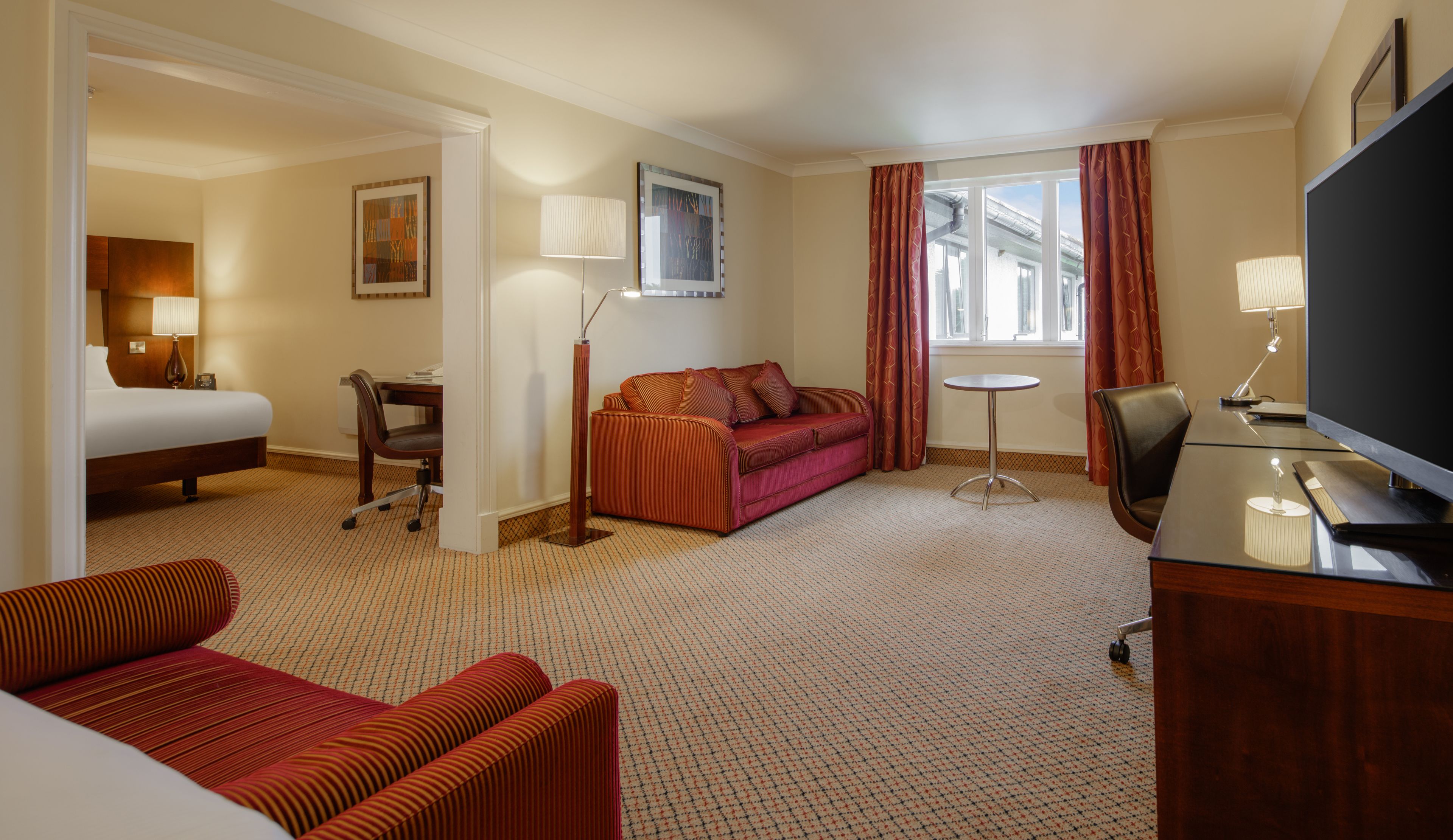 Quarto de hotel com cama, sofá, escrivaninha com cadeira, televisão e janela com cortinas.