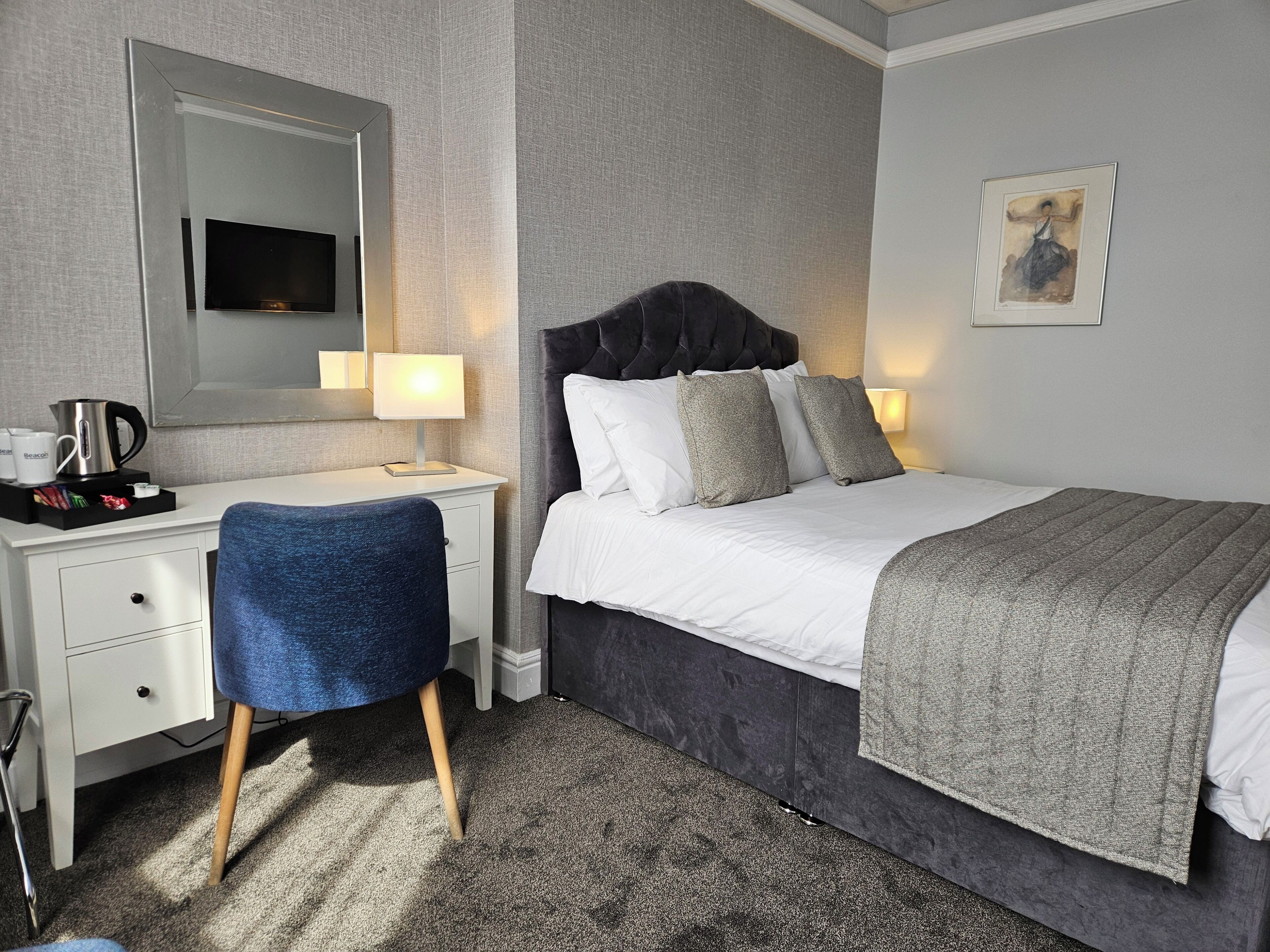 Habitación de hotel con cama, escritorio, silla, mesita de noche con lámpara, cafetera e imagen enmarcada en la pared.
