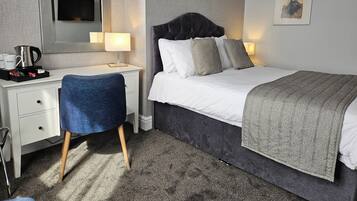 Habitación de hotel con cama, escritorio, silla, mesita de noche con lámpara, cafetera e imagen enmarcada en la pared.