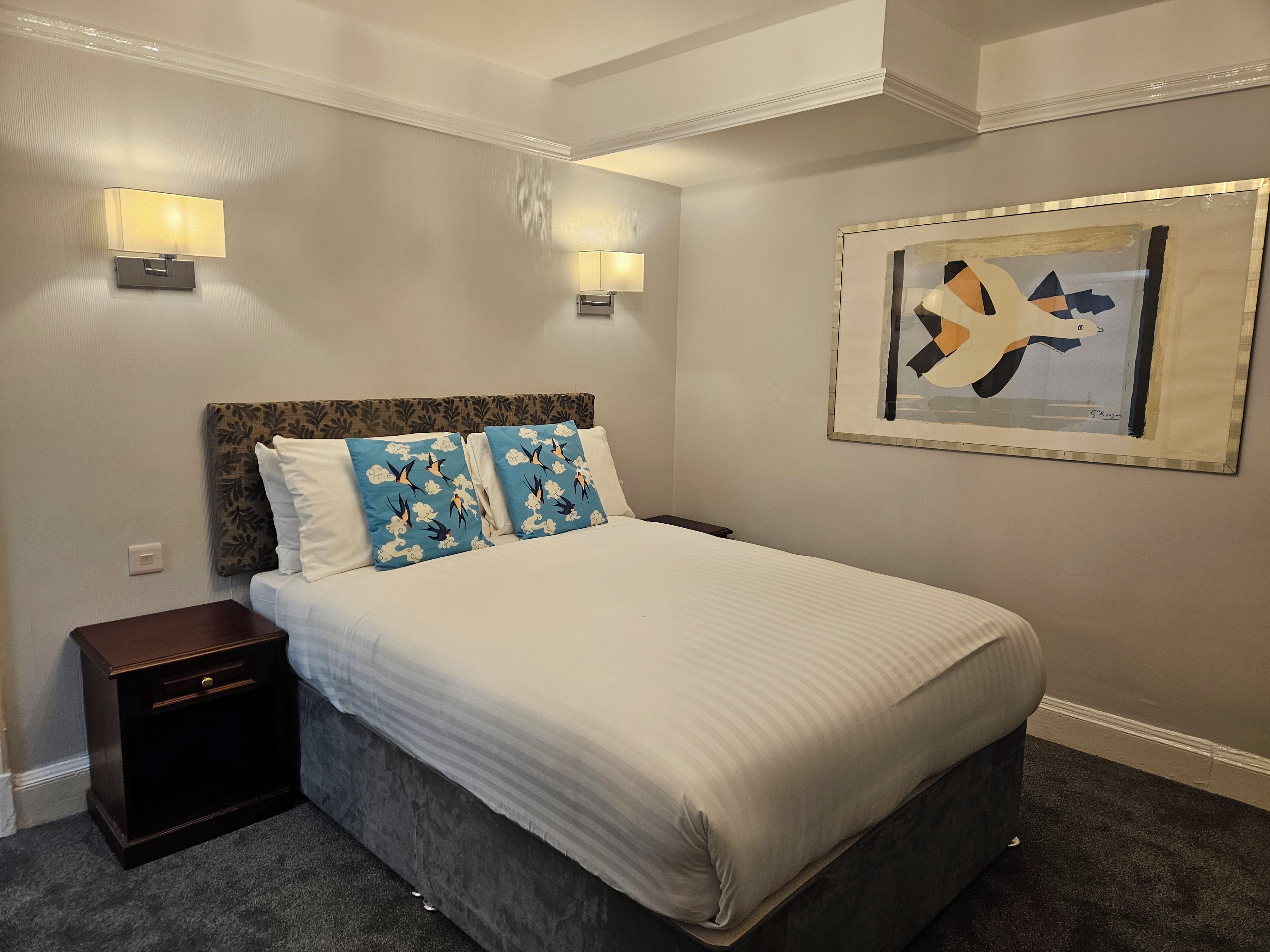 Ein Hotelzimmer mit einem Bett, zwei Wandlampen, einem Nachttisch, einem gerahmten Kunstwerk und einem Teppichboden.