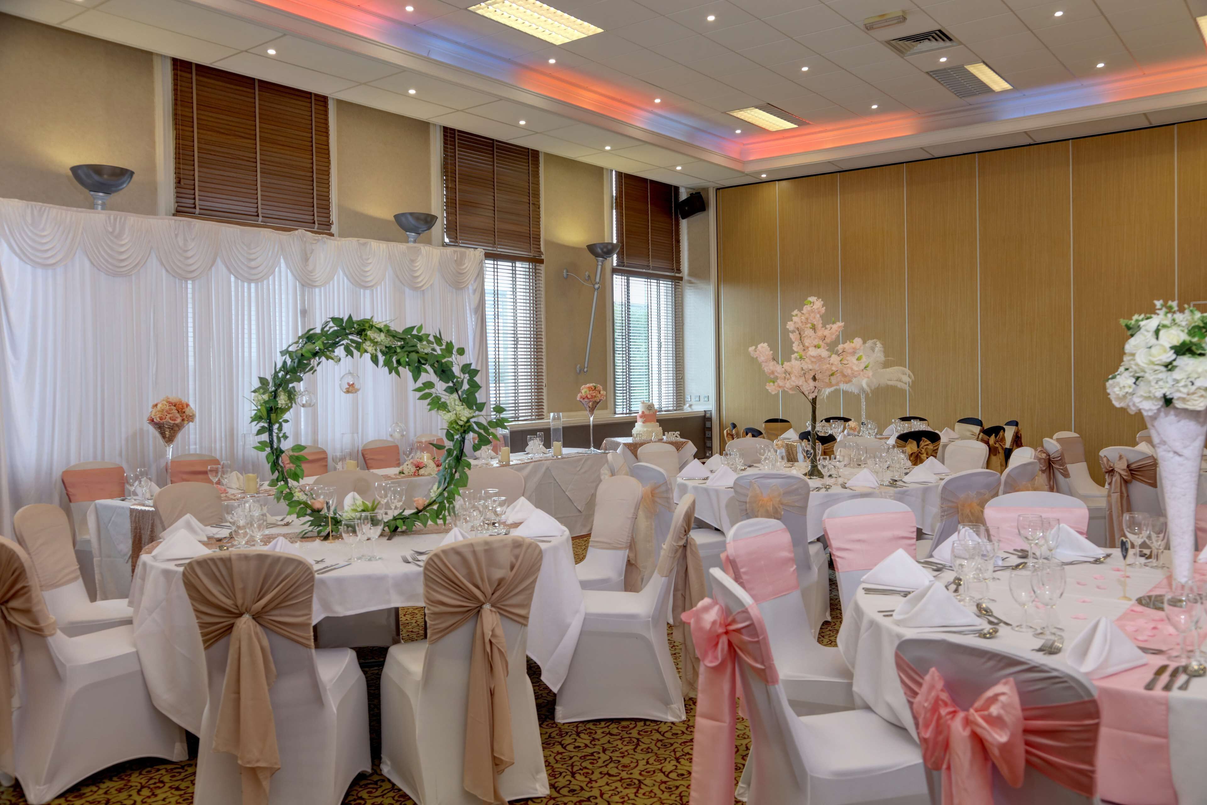 Un salón de banquetes con mesas redondas, sillas blancas y rosas, y centros de mesa florales.