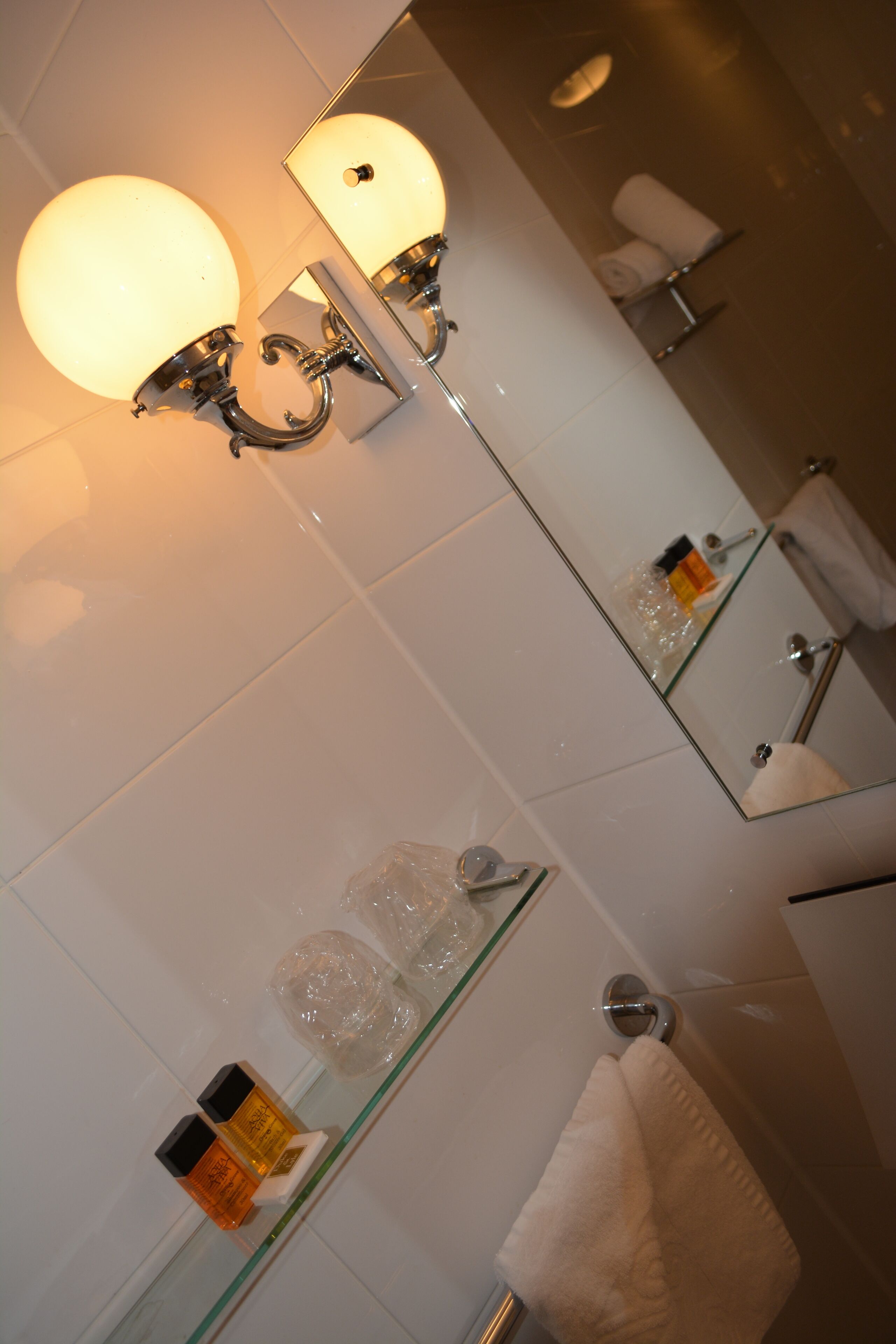 Un baño con repisa de vidrio que sostiene artículos de aseo, un perchero y un espejo con lámpara.